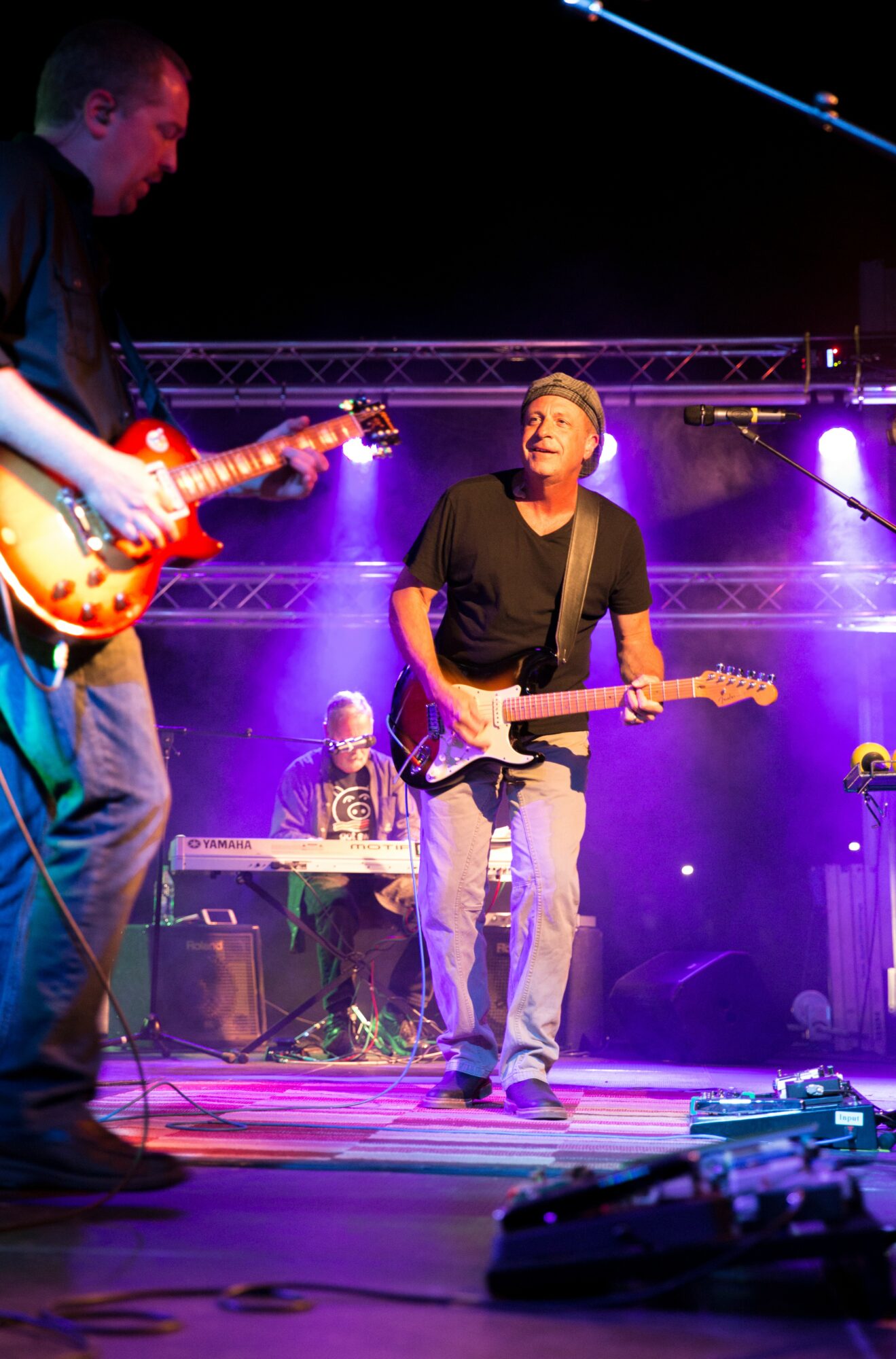 Three musicians perform on stage with purple lighting, one playing guitar, another on keyboard, and a third singing.