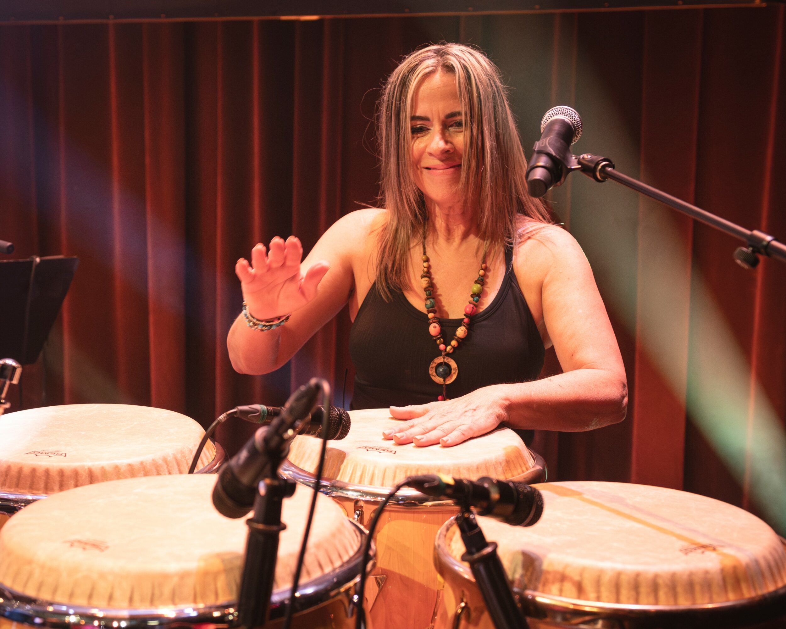 Woman playing drums on stage with red curtains, smiling, wearing a black top and necklace, microphone nearby.