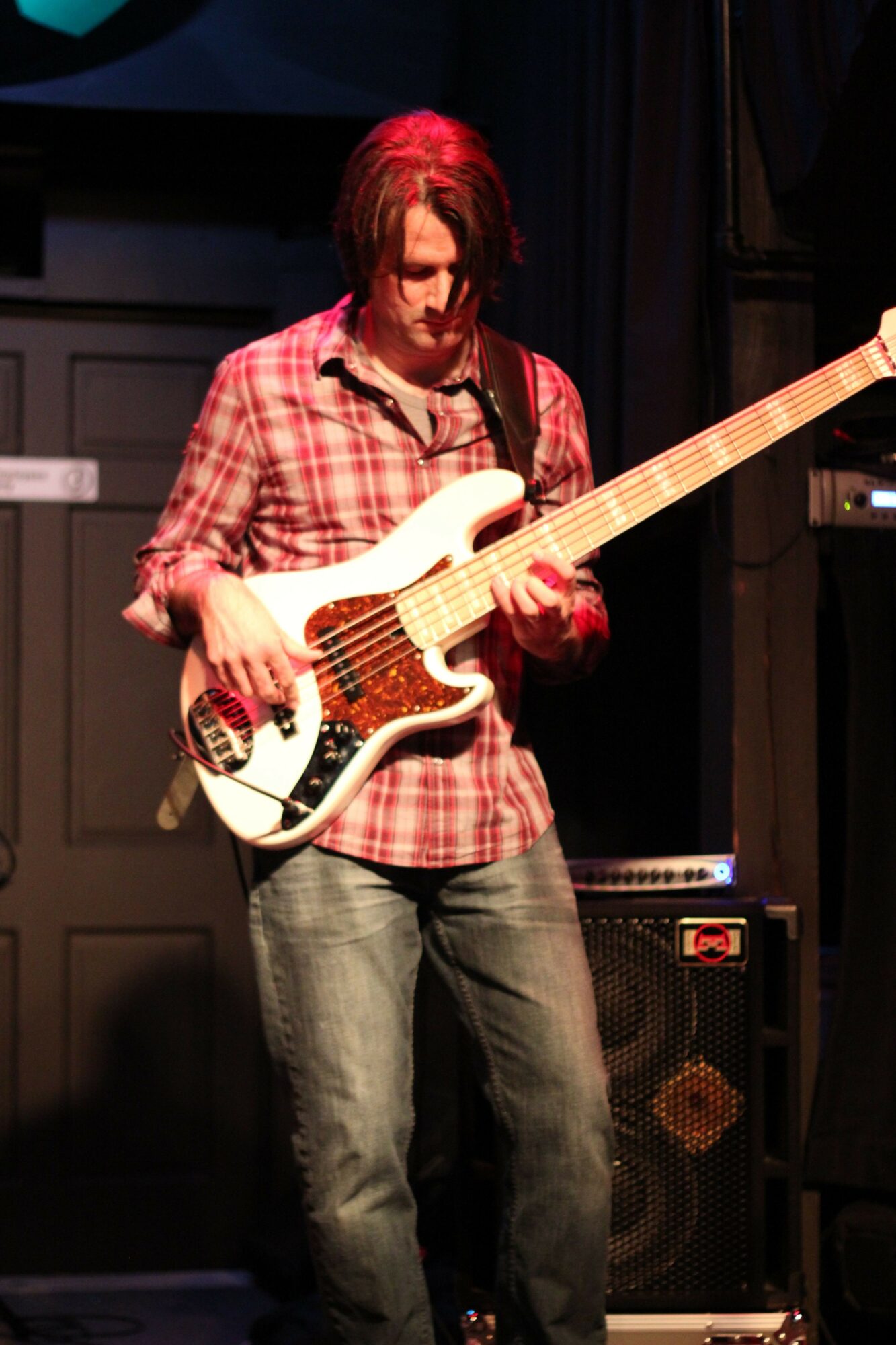 Person with long hair wearing a red plaid shirt holding an electric guitar on stage.