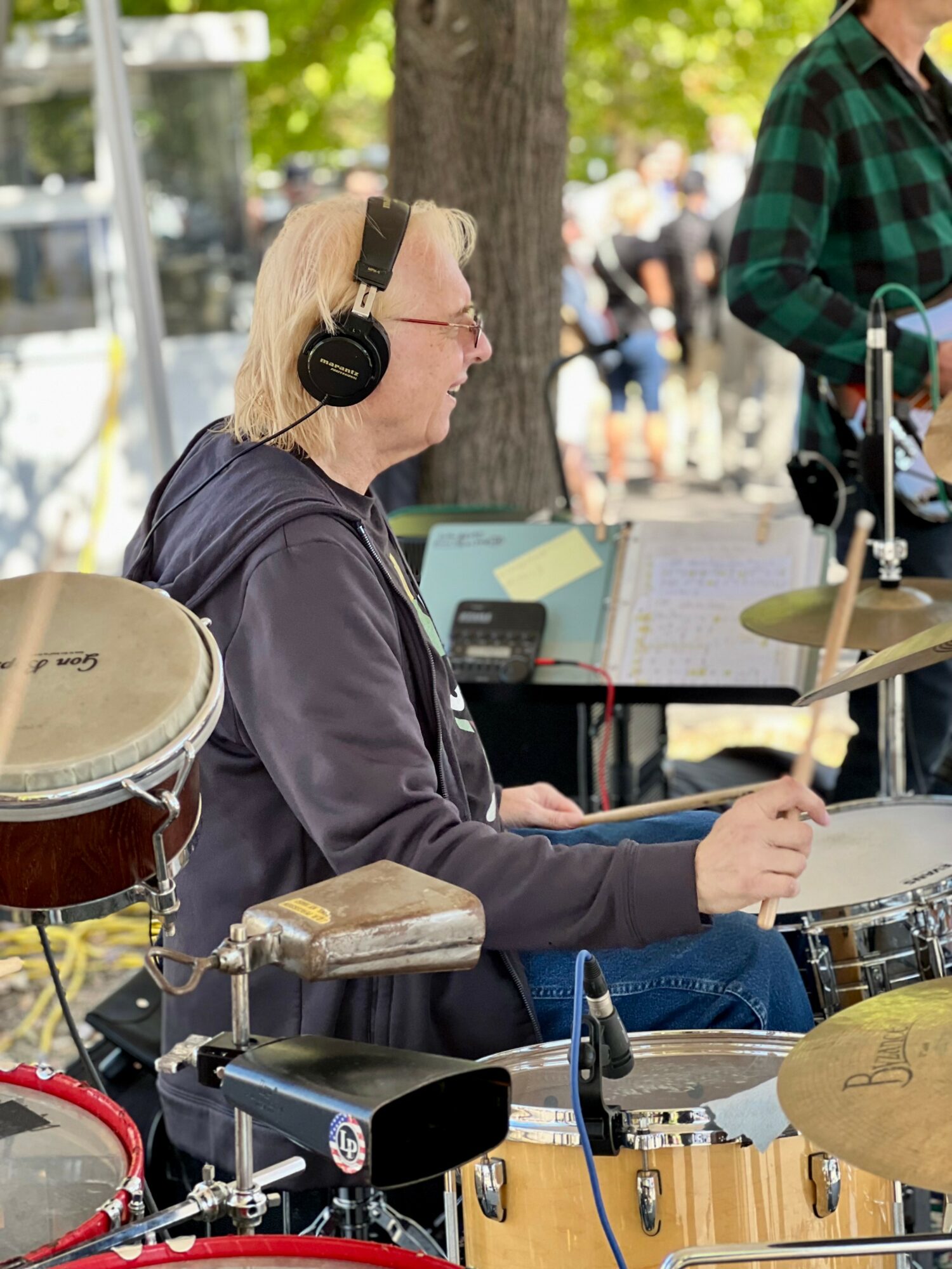 A woman with blonde hair wearing headphones plays drums outdoors, with trees and people in the background.
