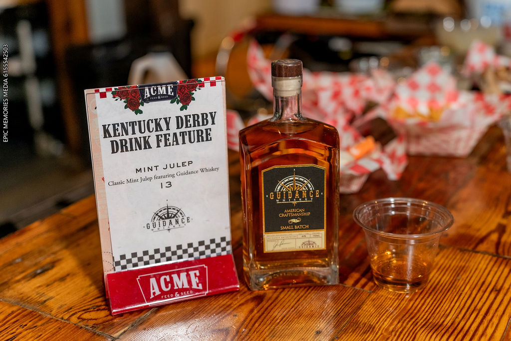 Bottle of whiskey, plastic cup, and Kentucky Derby drink feature sign on wooden table, blurred background with flowers.