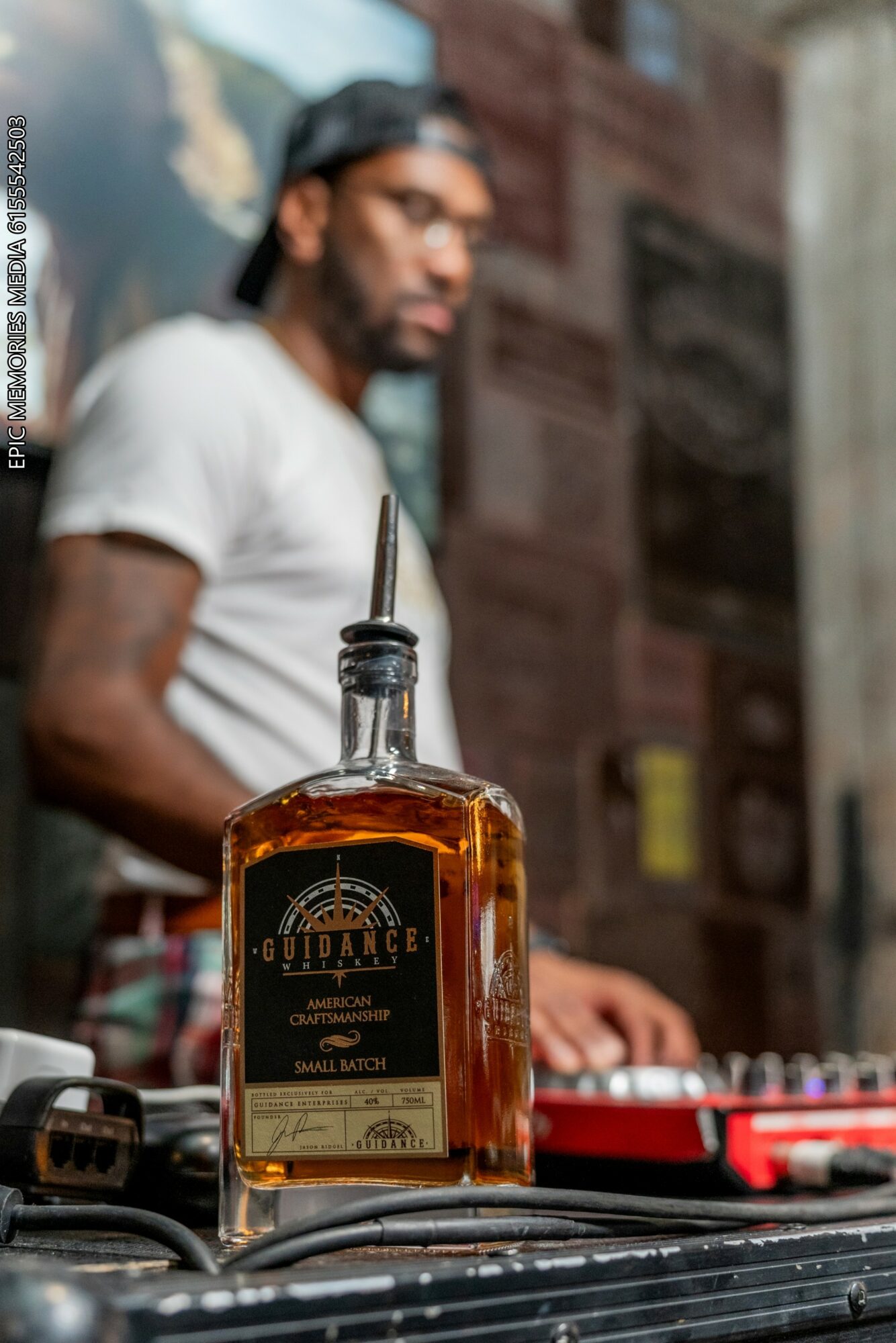 A bottle of liquor in focus with a blurred person in the background, outdoors setting, wooden wall behind.