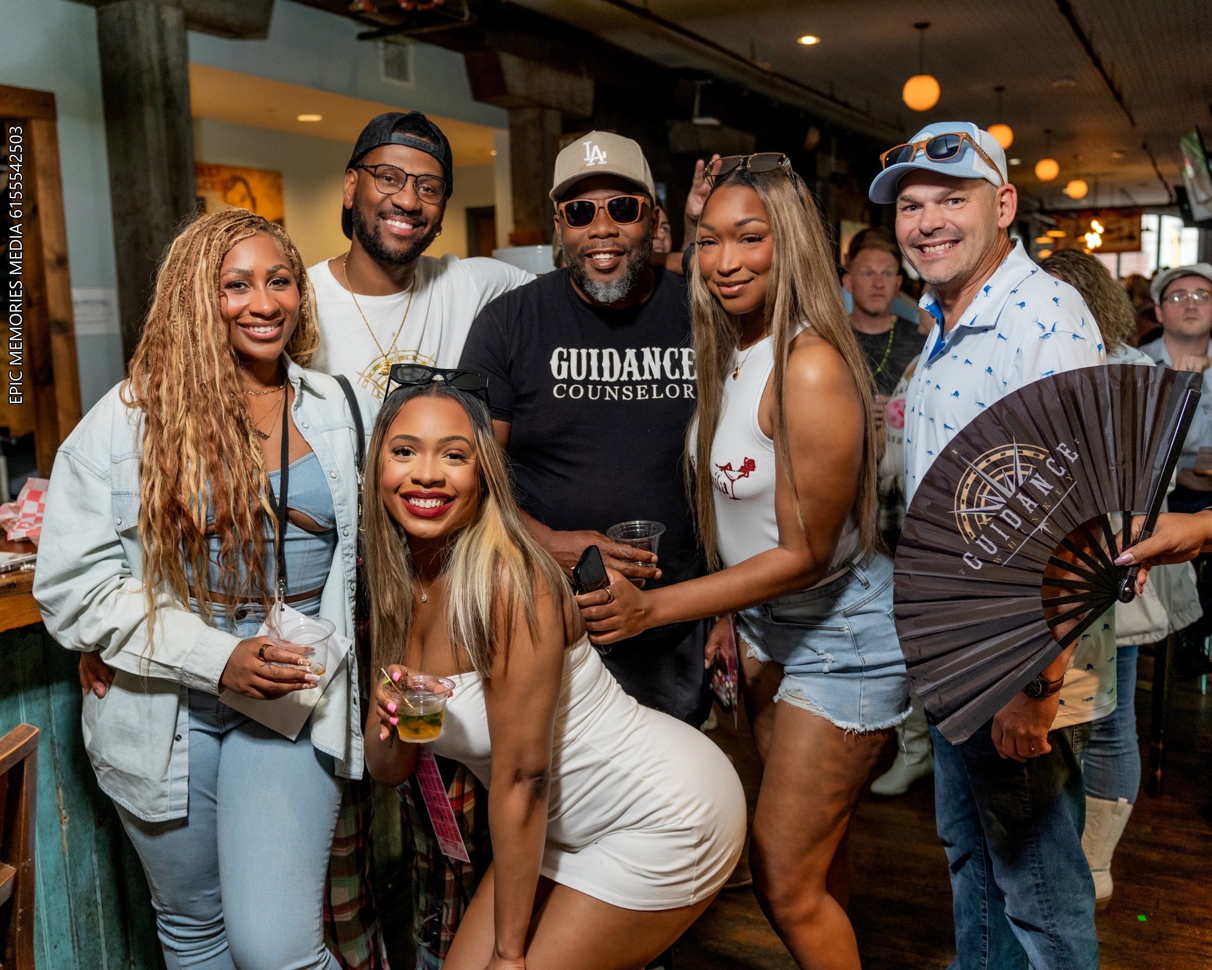 Group of six diverse people smiling and posing together indoors, some holding drinks, in casual attire.