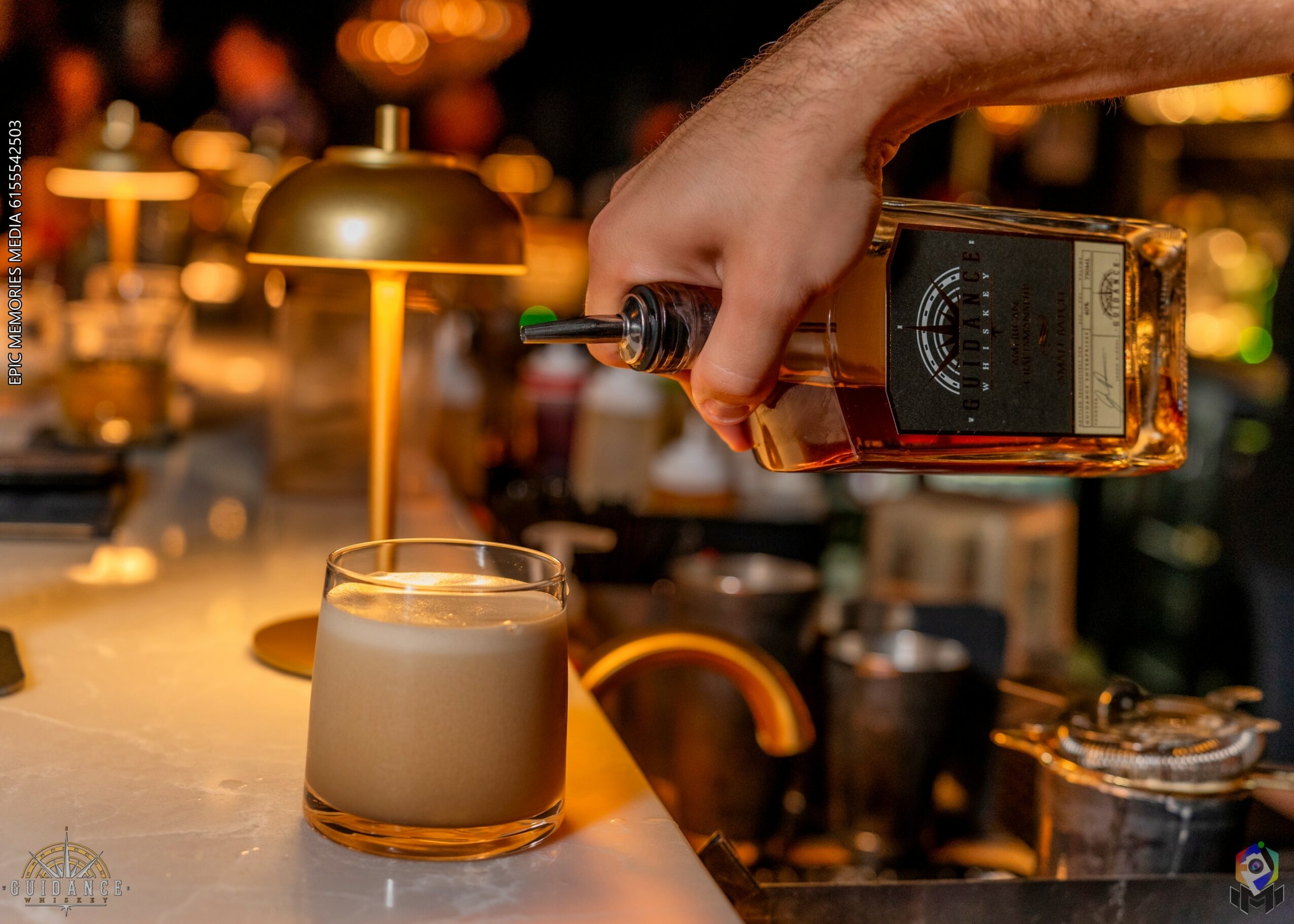 Person pouring whiskey into a glass with a bar background, warm lighting, and a modern lamp nearby.