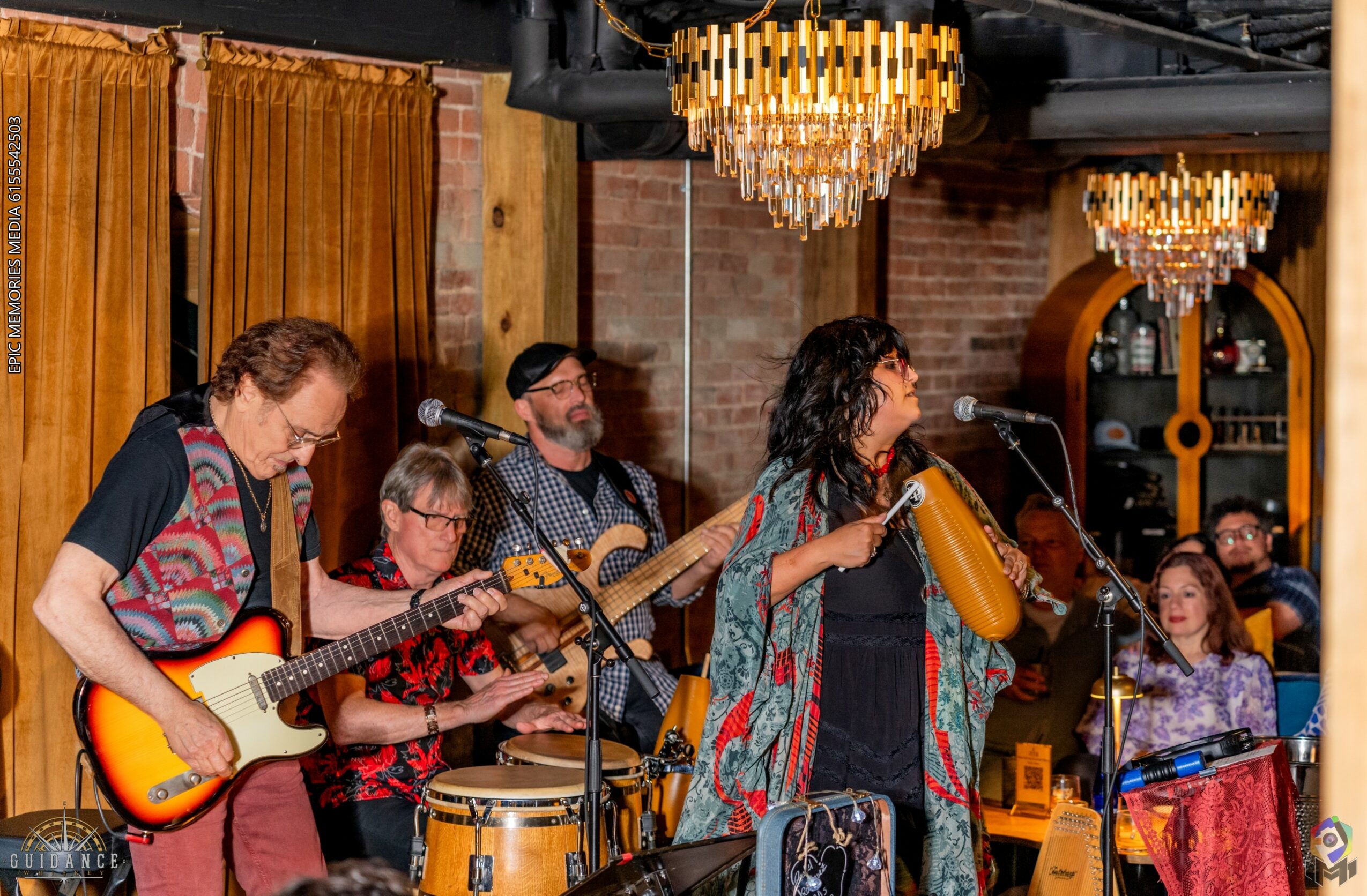 Group of musicians performing with guitars, drums, and a microphone in a warmly lit venue with chandeliers and wooden decor.