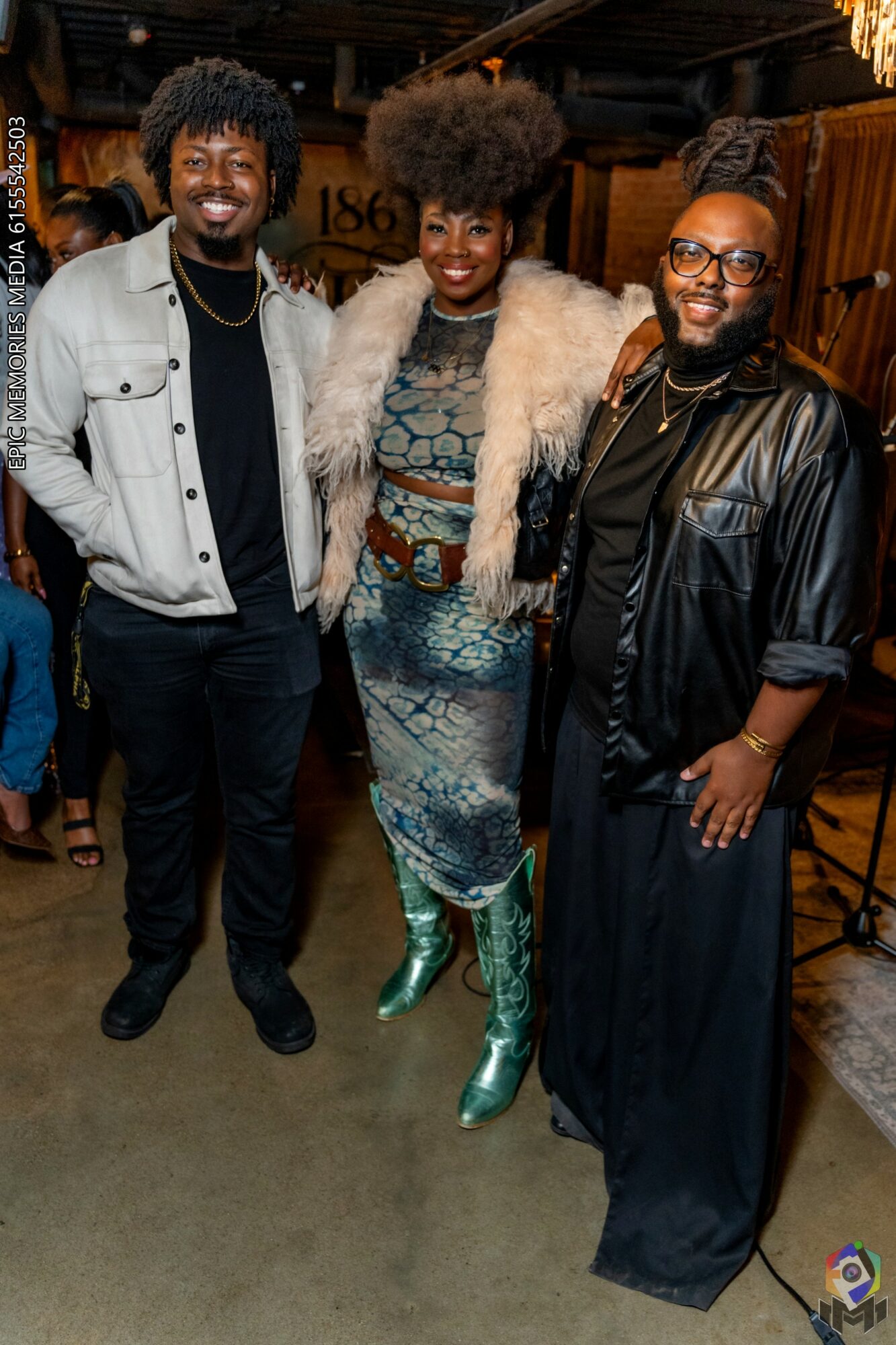 Three people smiling and posing together indoors, with a woman in the center wearing a patterned dress and green boots.
