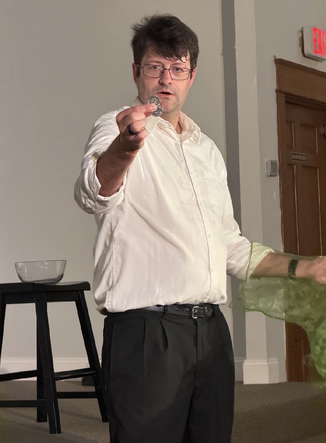 Man with glasses and dark hair pointing forward, wearing a white shirt and black pants, standing indoors.