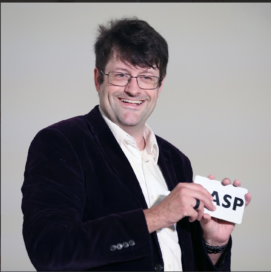 Man with glasses and dark hair smiling, holding a white mug with 'ASP' written on it, wearing a dark blazer and white shirt.