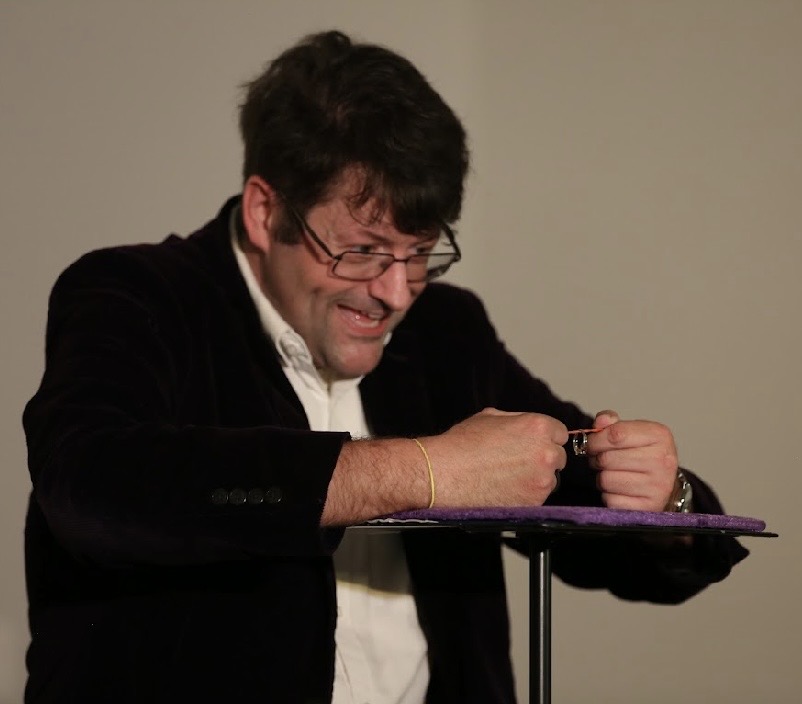 Person smiling, wearing glasses, holding a pen, with a purple object on a stand, against a plain background.