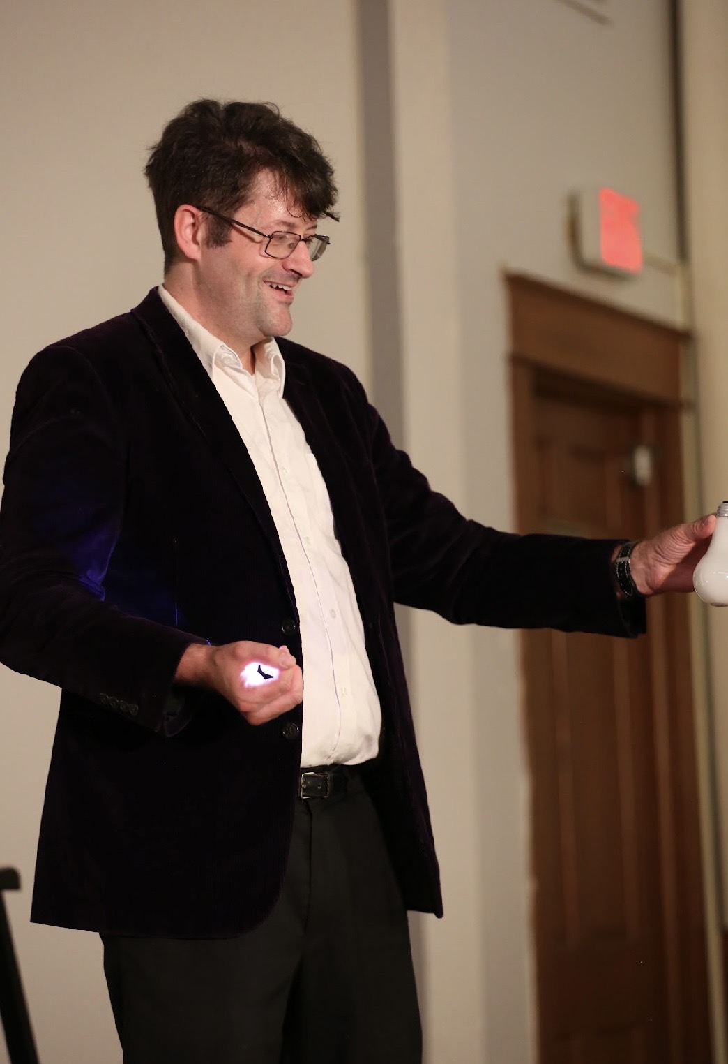Man with glasses smiling, wearing a black blazer and white shirt, standing indoors with a door in the background.