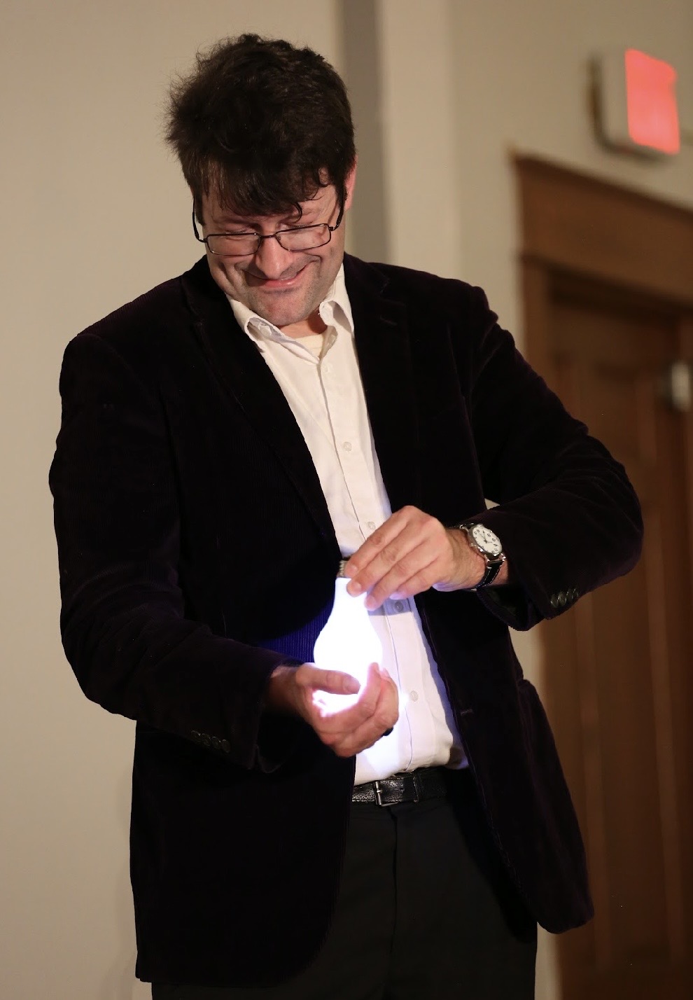 Man with glasses and dark hair looking down, holding a glowing object in his hands, wearing a black blazer and white shirt.