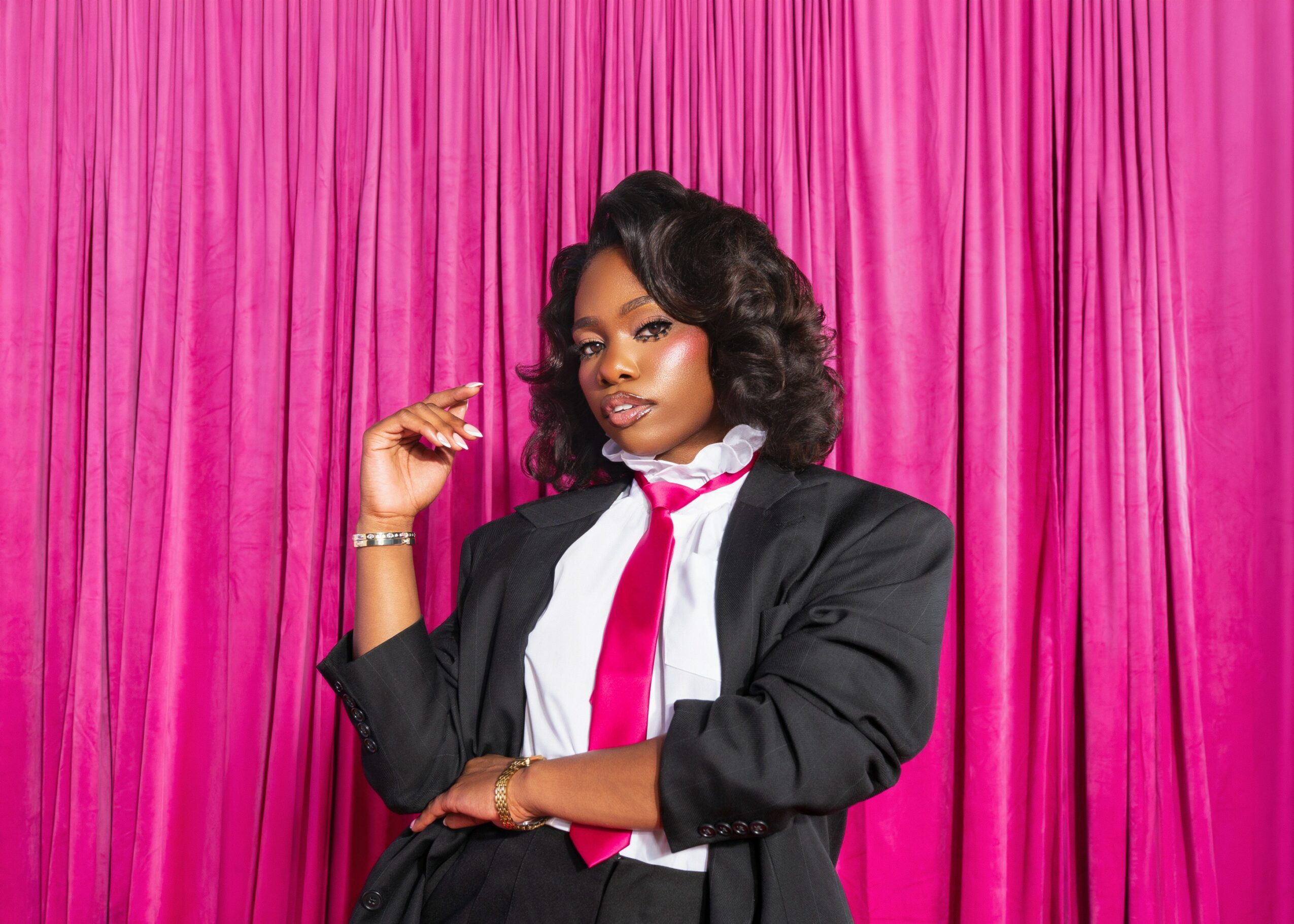Woman with styled hair and makeup, wearing a black blazer, white shirt, and pink tie, standing in front of pink curtains.
