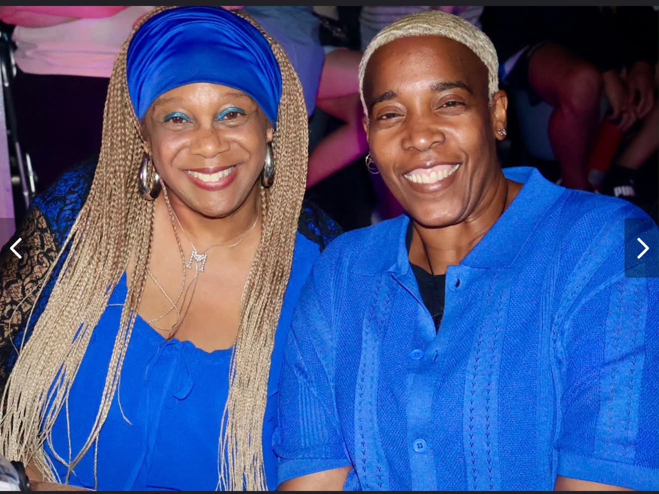 Two smiling women in blue outfits, one with long braided hair and a blue headwrap, the other with short blonde hair.