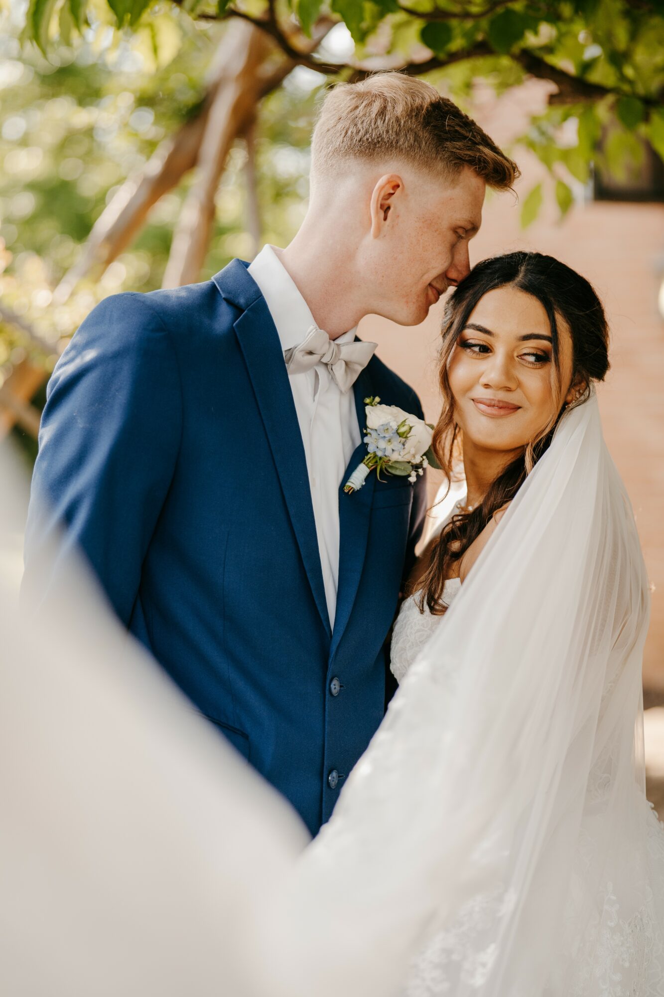 A bride and groom stand close outdoors, with trees and sunlight in the background, smiling softly at each other.