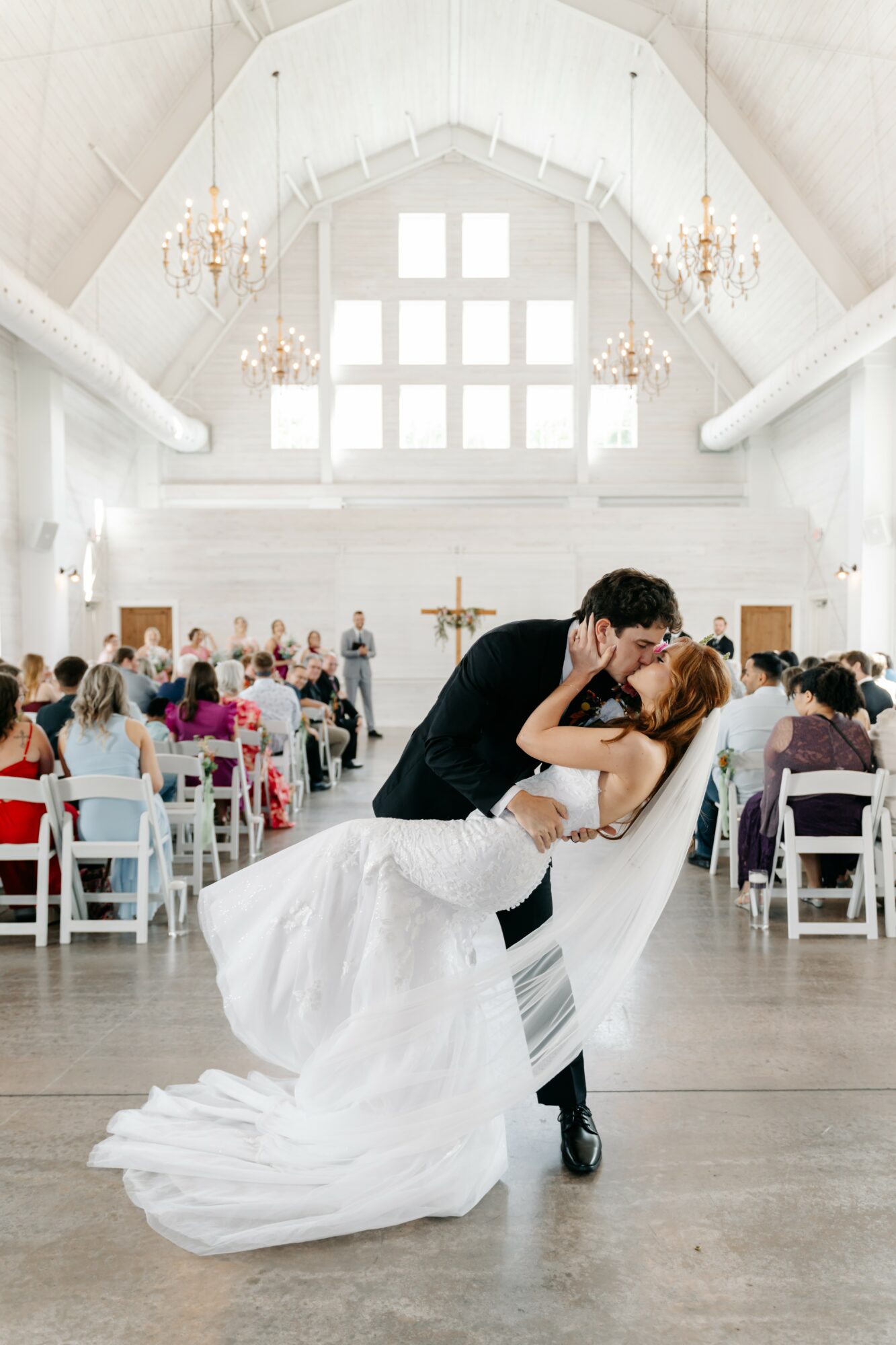 A couple dances in a bright, elegant wedding venue with high ceilings and chandeliers, surrounded by seated guests.