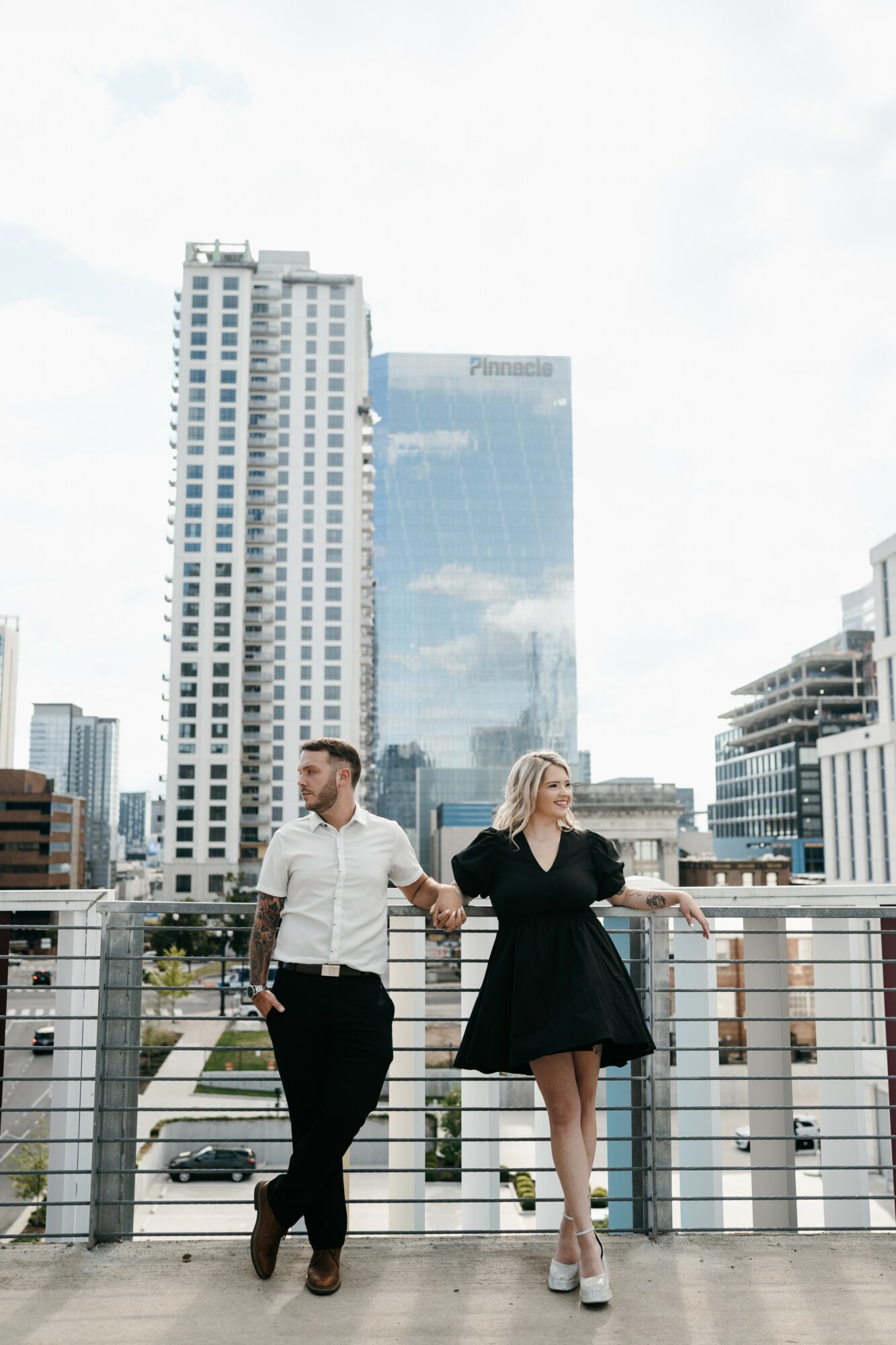 A man and woman walk on a rooftop with a city skyline in the background, holding hands and looking at each other.