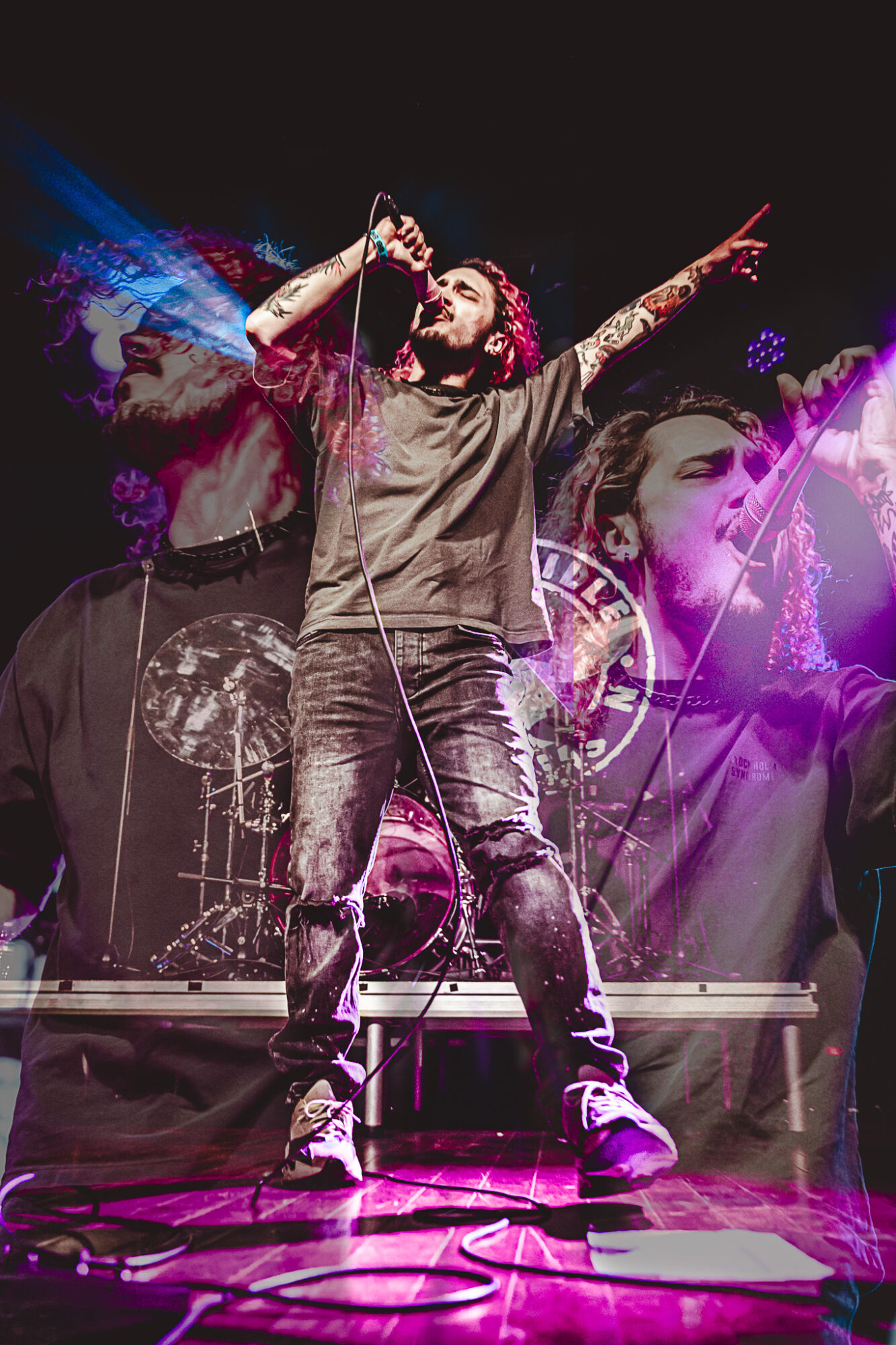 Male singer with tattoos performs on stage with a microphone, colorful background, and a large face image behind him.