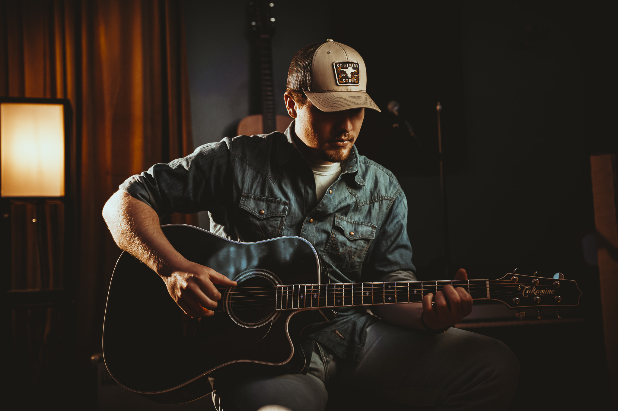 Person playing guitar in a dimly lit room with warm lighting, wearing a cap and casual clothing.