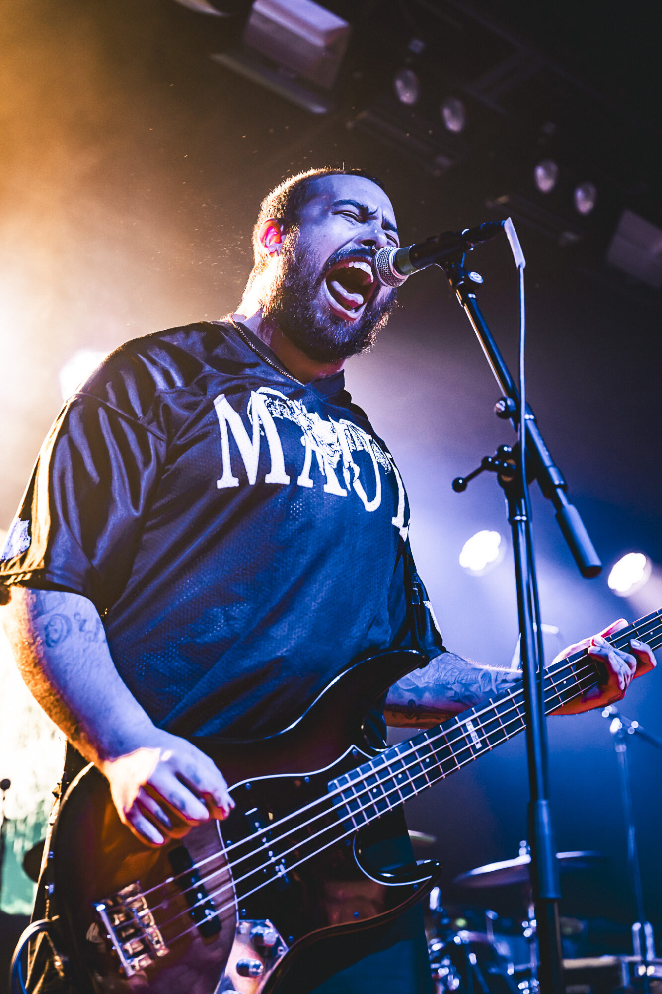 Musician passionately singing into microphone while playing bass guitar on stage with colorful lighting.