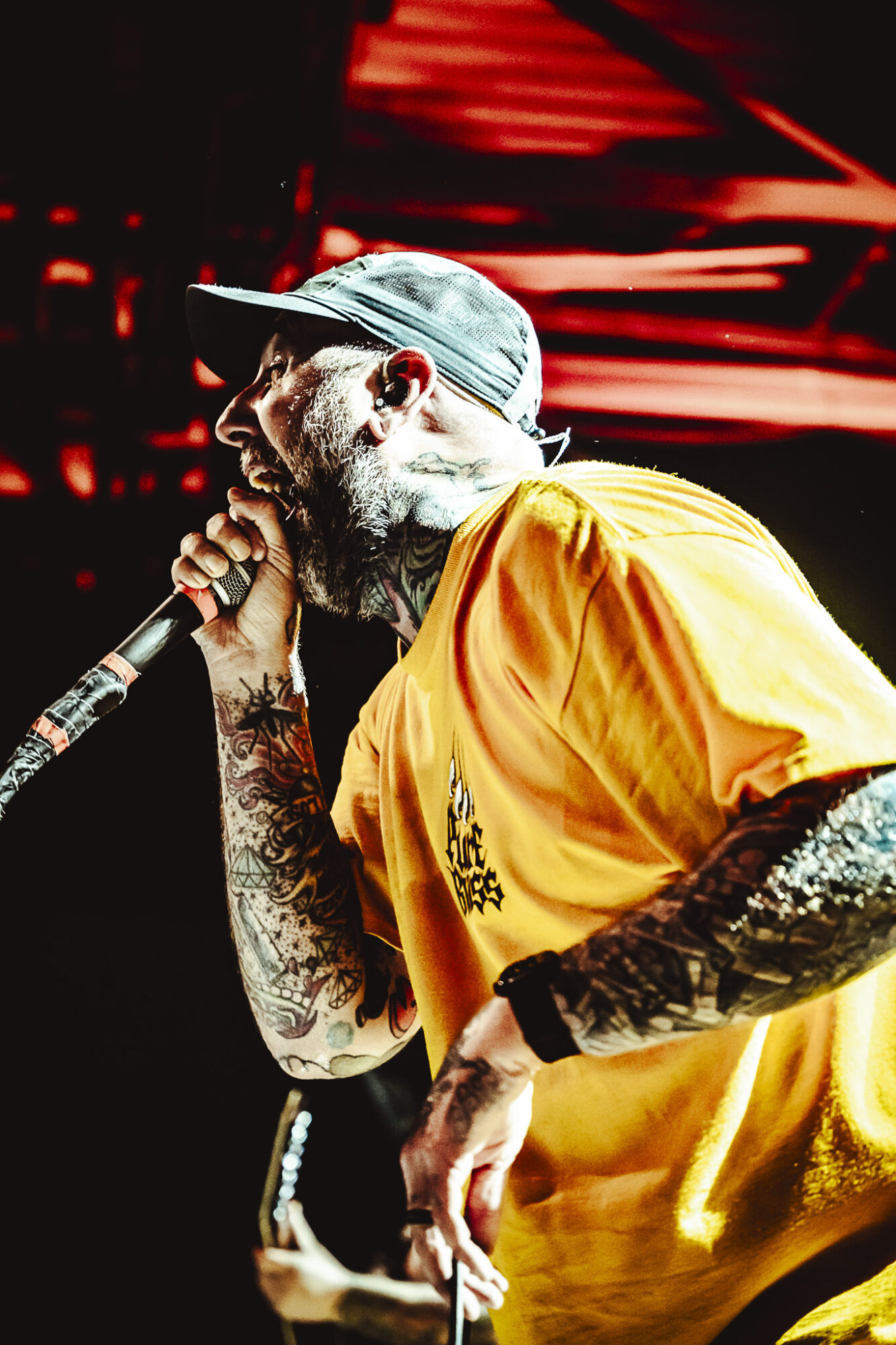 Male performer singing into a microphone, wearing a cap and yellow shirt, with tattoos on arms, on stage with red lighting.