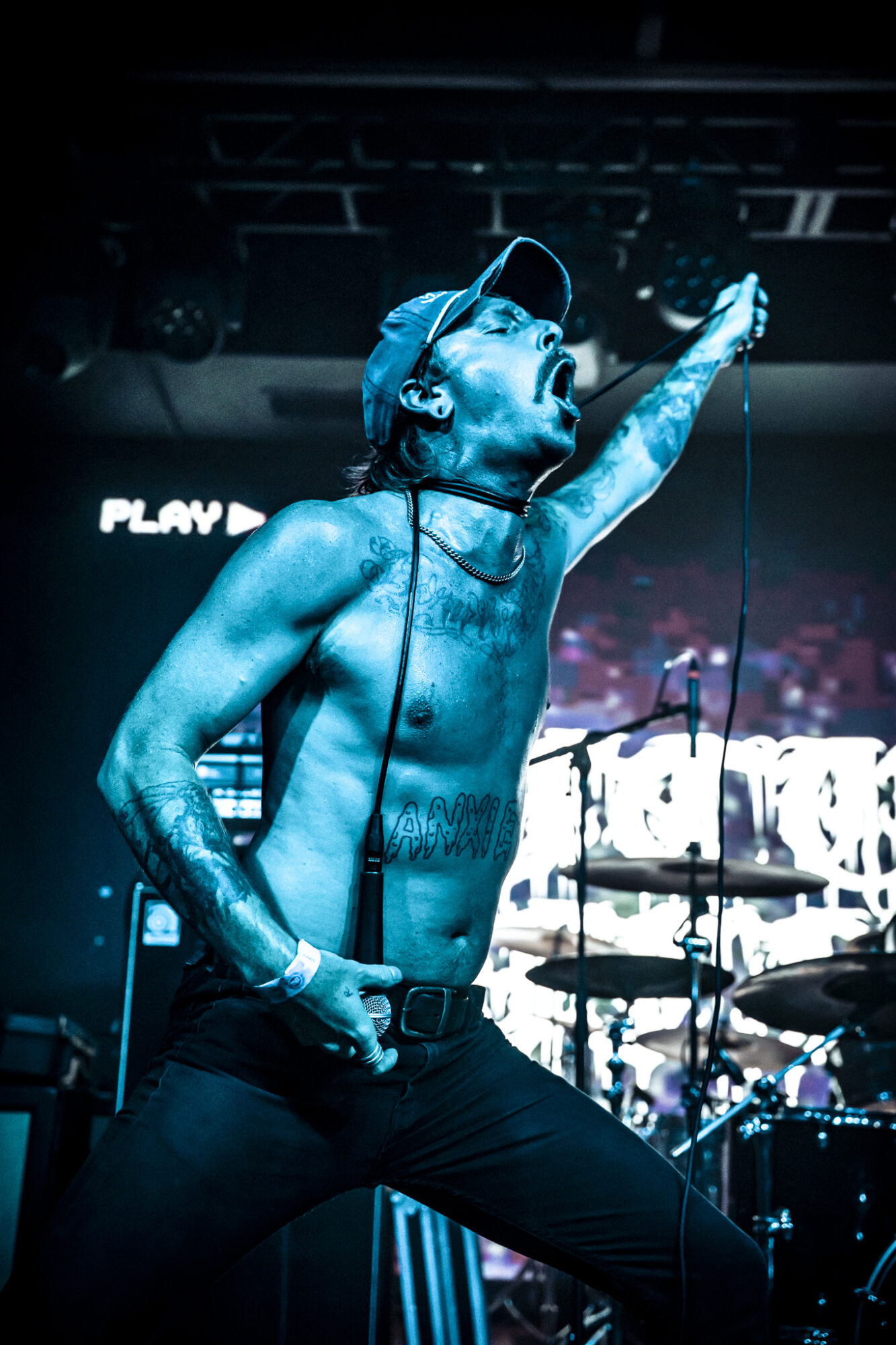 Male performer singing passionately on stage, holding a microphone, with drums in the background, blue lighting, shirtless, wearing a cap.
