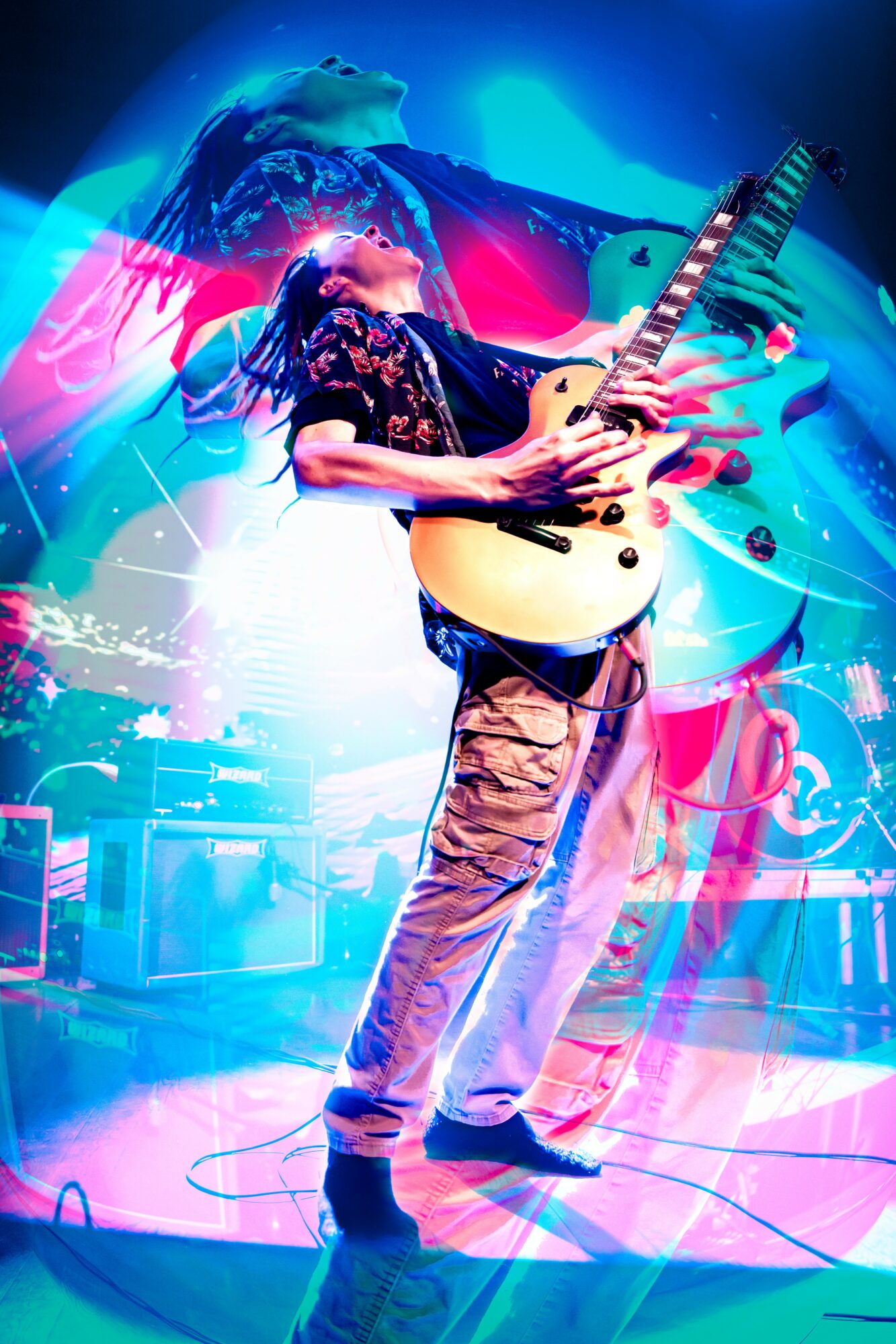 Person playing electric guitar on stage with colorful lights and abstract background, wearing patterned shirt and jeans.