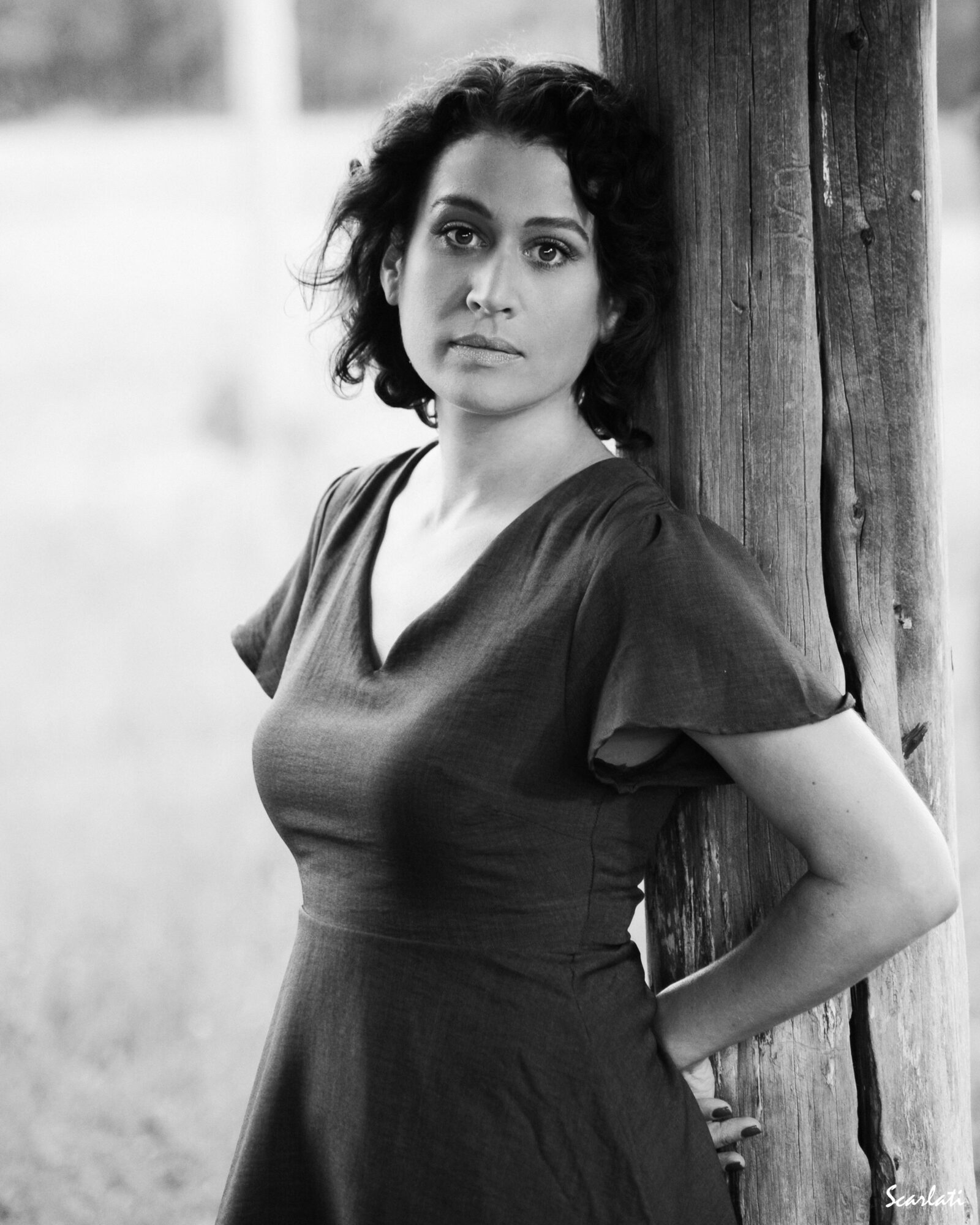 Woman with short curly hair leaning against a wooden post outdoors, wearing a dark dress, looking at the camera.