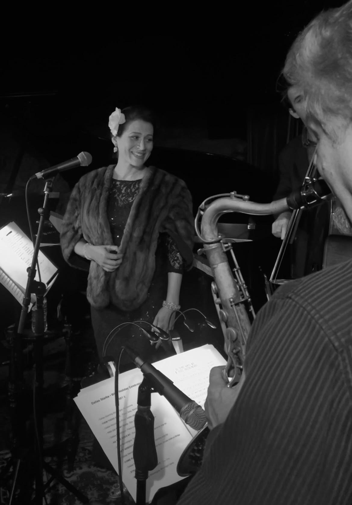 A woman stands on stage with a microphone, smiling, while a person plays a saxophone nearby, with music sheets visible.