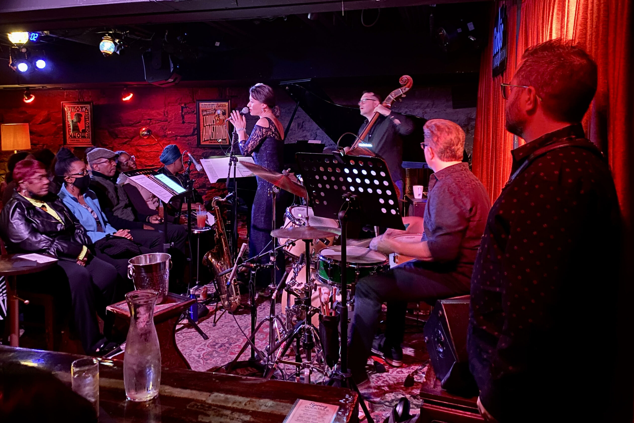 Musicians perform on stage with an audience watching in a dimly lit venue, red curtains and musical instruments visible.