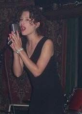 Woman in black dress holding a phone, smiling, with dark background and patterned wall.