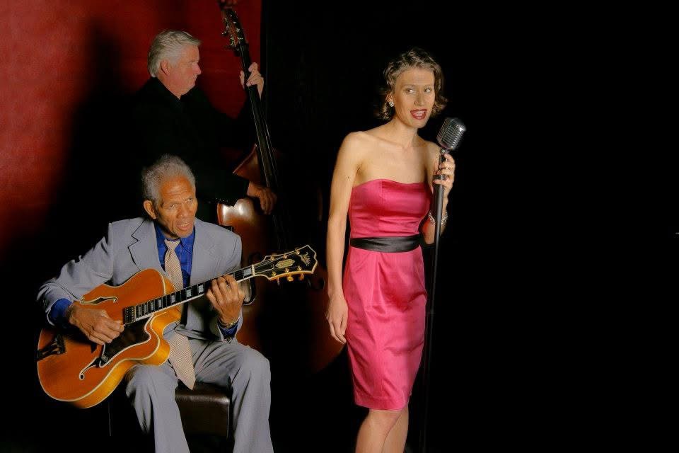 A woman in a pink dress holding a microphone, with two men playing instruments behind her, against a dark background.