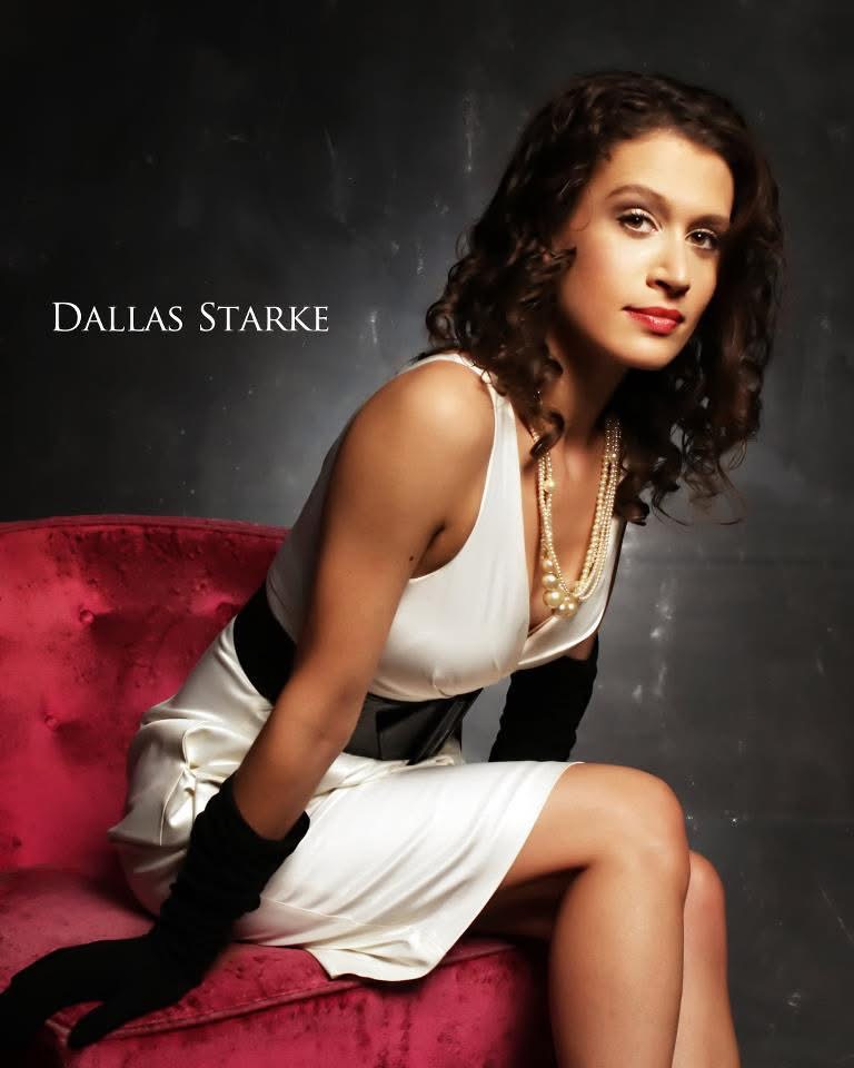 Woman with curly hair in a white dress sitting on a red chair, looking at the camera, dark background.