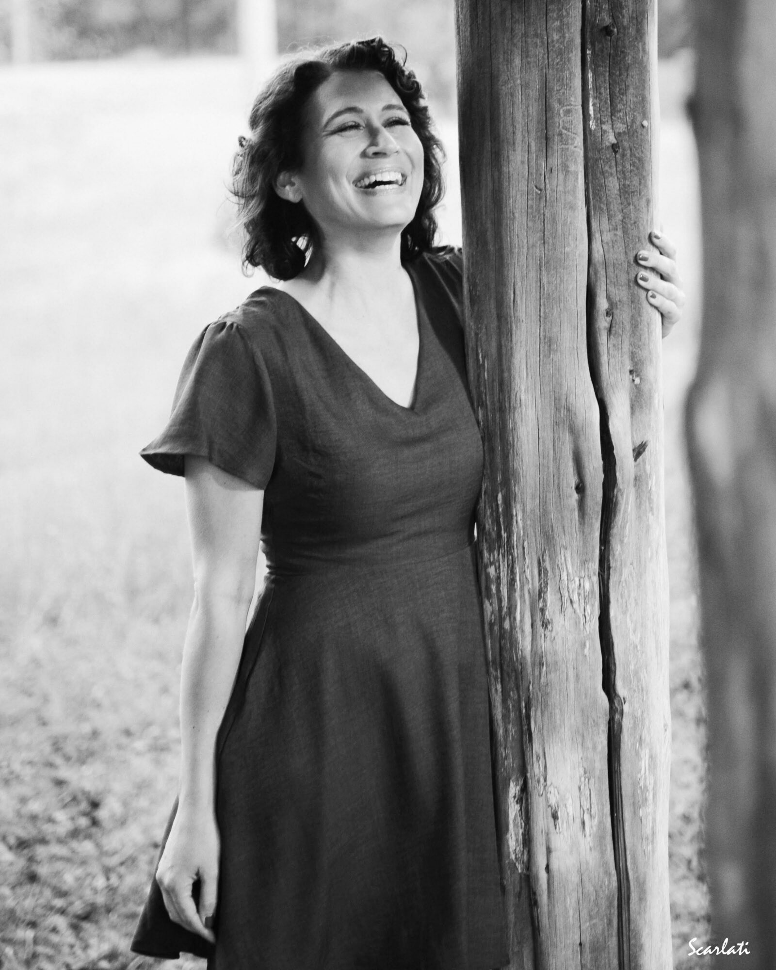 Woman smiling and laughing outdoors, holding a tree trunk, wearing a dark dress, in black and white.