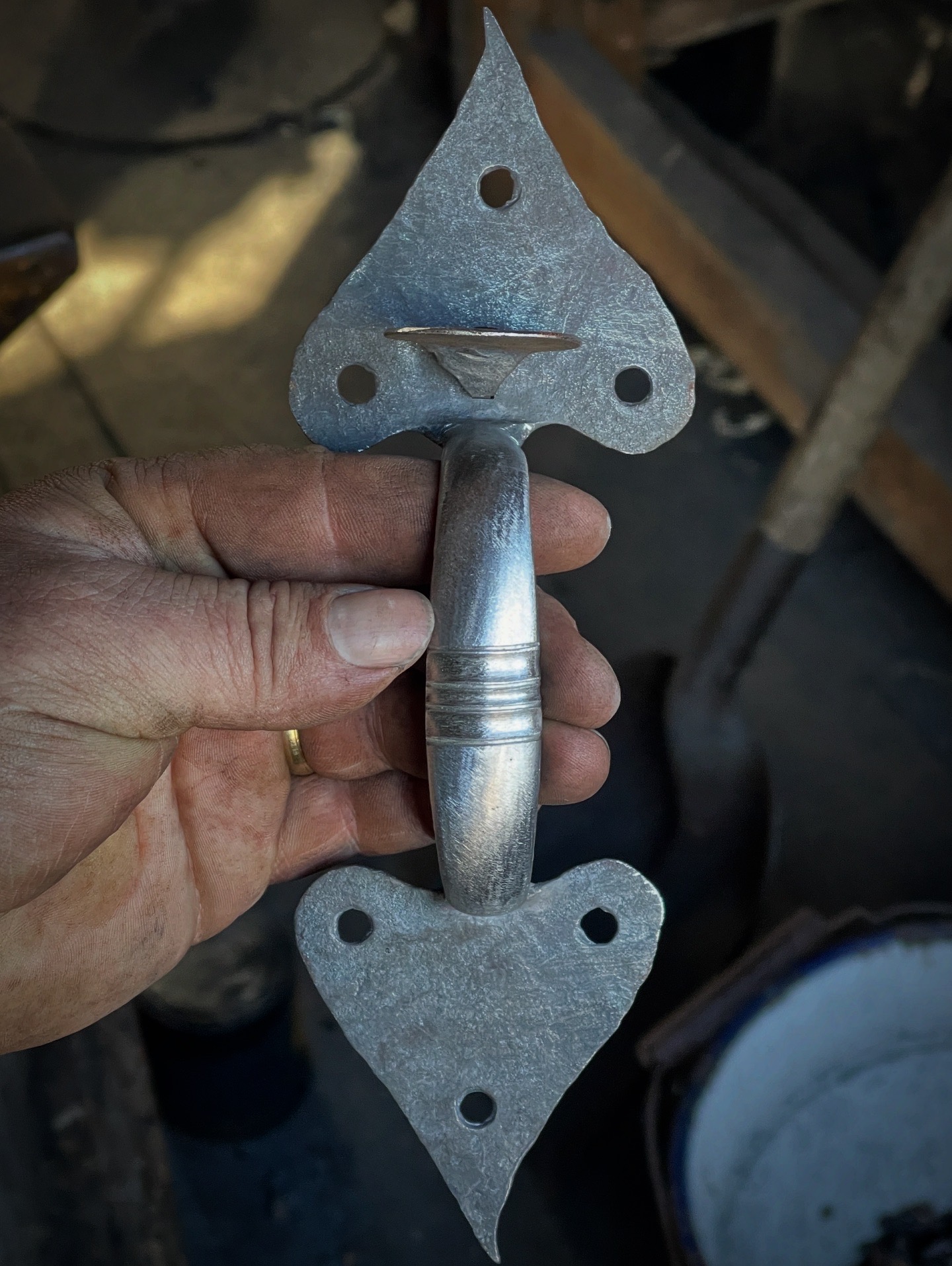 Hand holding a metal tool with two heart-shaped ends and a central handle, in a workshop setting.