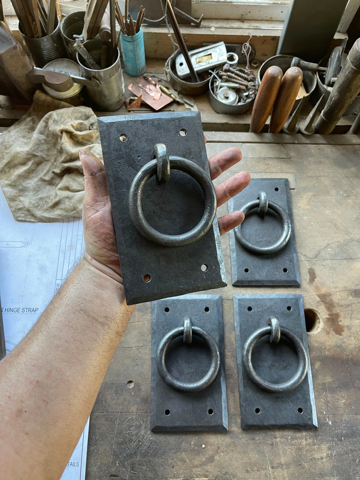 Hand holding a metal ring on a rectangular metal plate, with three similar plates on a wooden surface, workshop background.