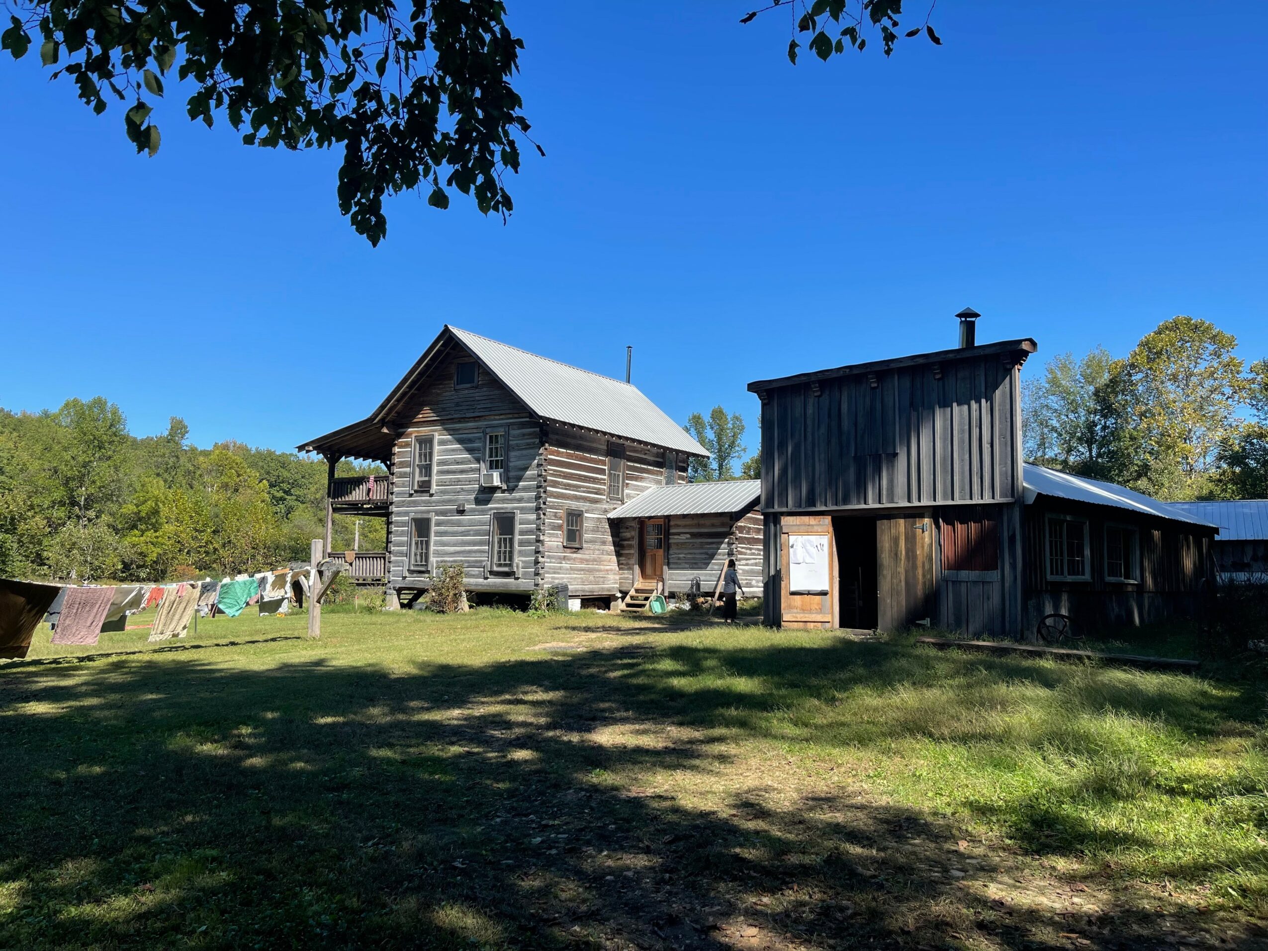 Two wooden buildings in a grassy area with trees and a clear blue sky, one larger and elevated, the other smaller and attached.