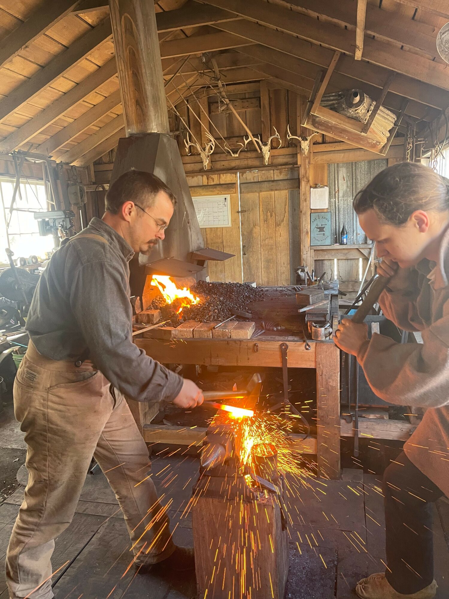Two people working with a welding machine in a workshop, sparks flying, wooden walls, and tools in the background.