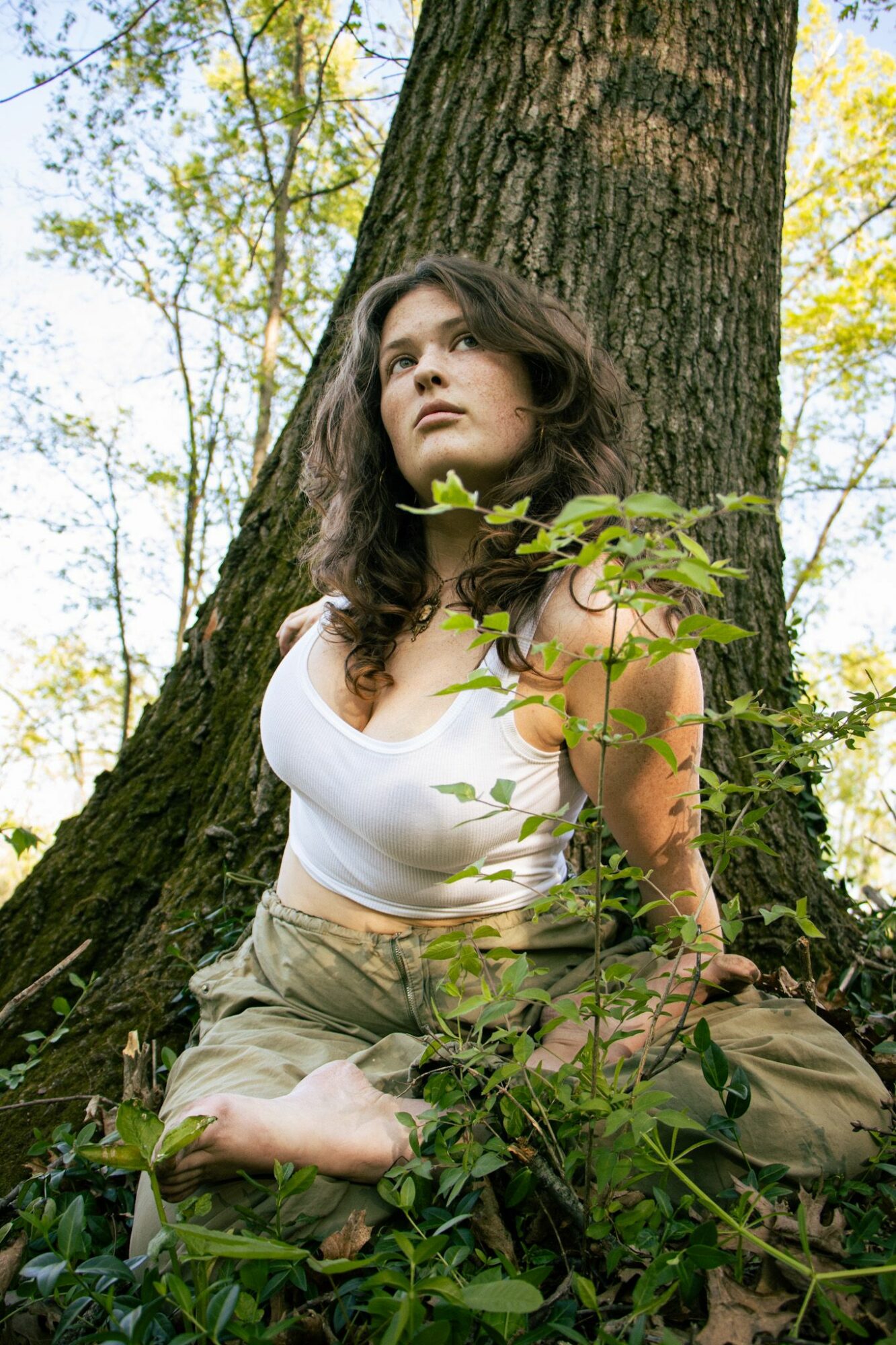 Woman sitting against a tree in a forest, surrounded by green leaves and sunlight, looking upward.