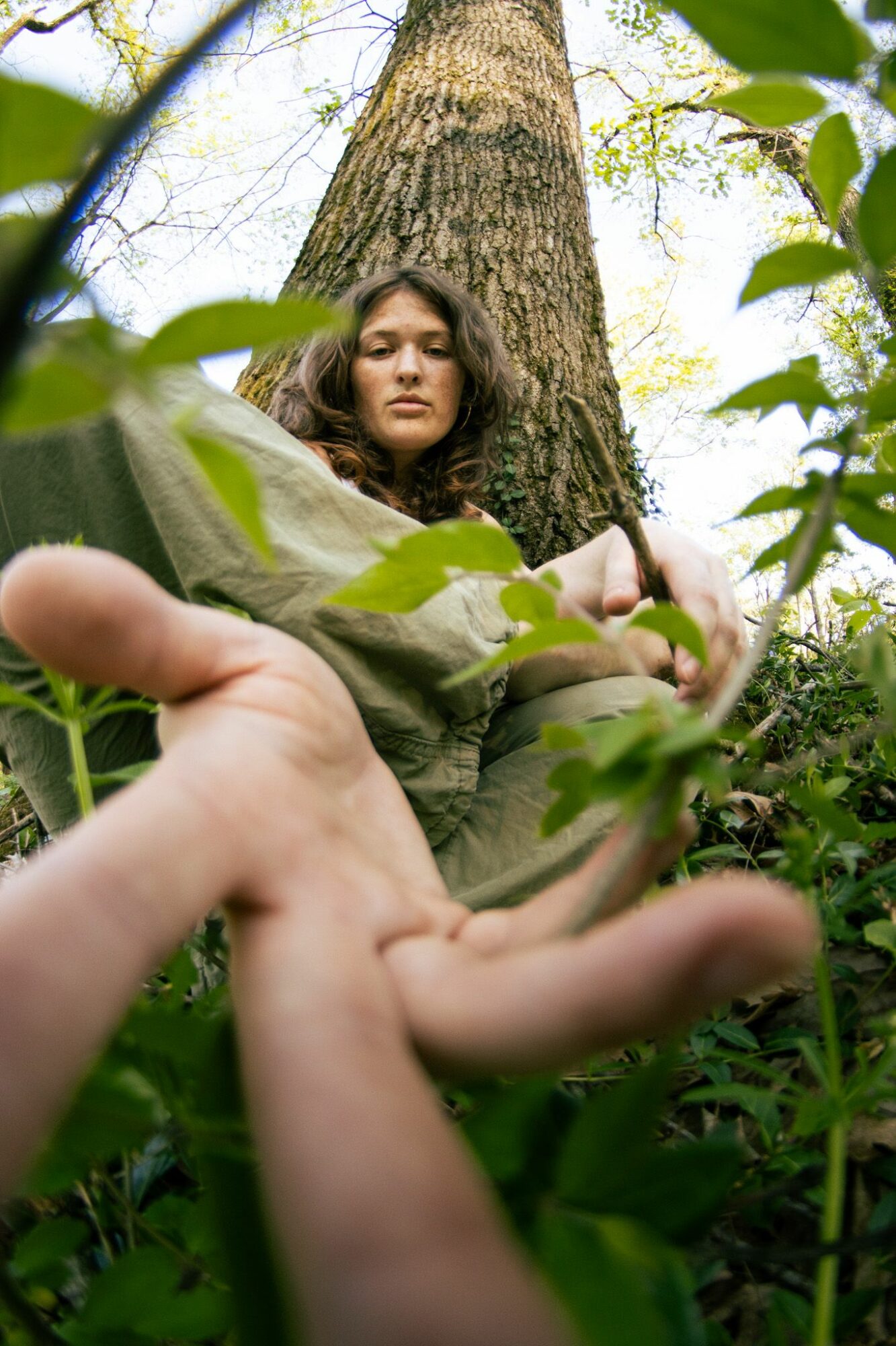 Person lying on ground with a large tree behind, surrounded by green leaves, looking at the camera.