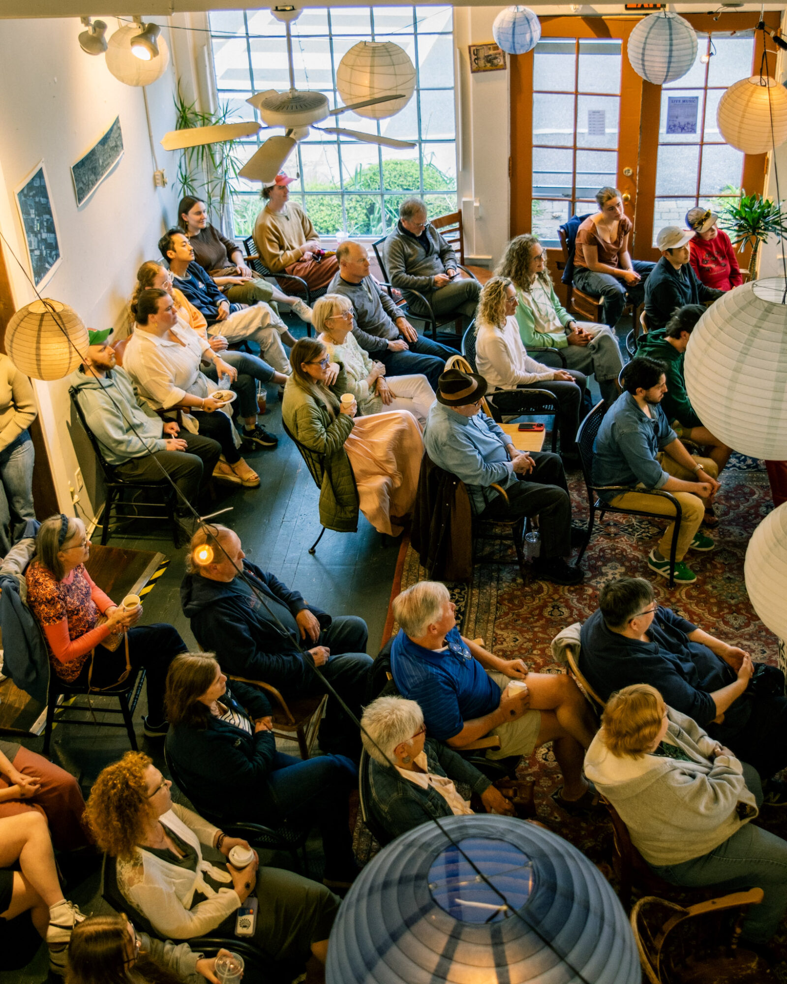 People seated in a room with large windows, hanging paper lanterns, and plants, attending a presentation or event.