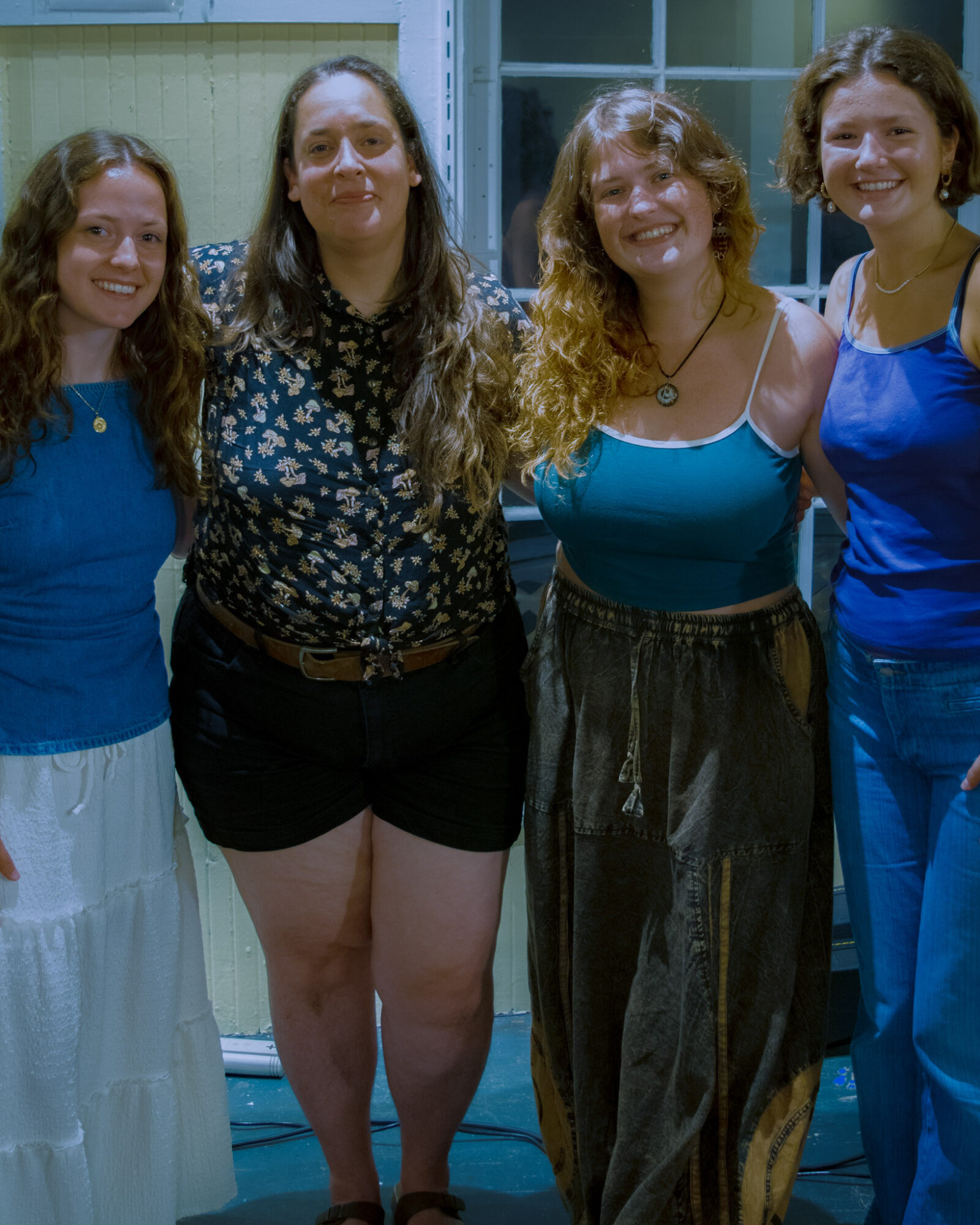 Four women standing together indoors, smiling, with a window and green wall in the background.