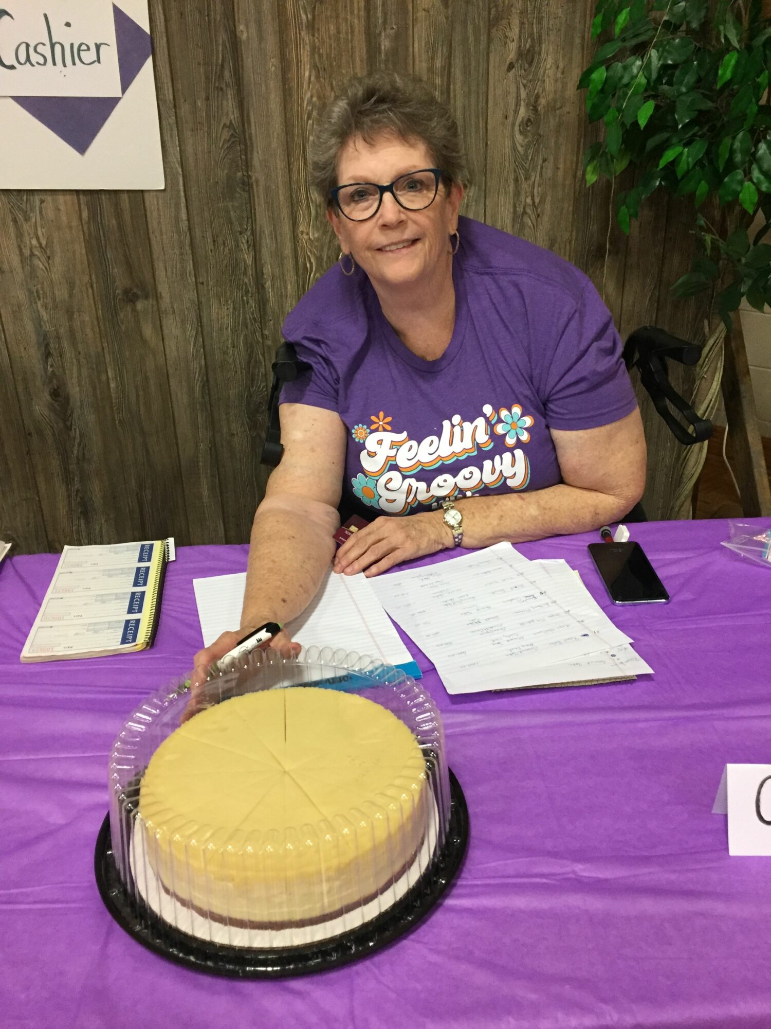 Woman with glasses and purple shirt sitting at table with a large yellow cake, writing on paper.