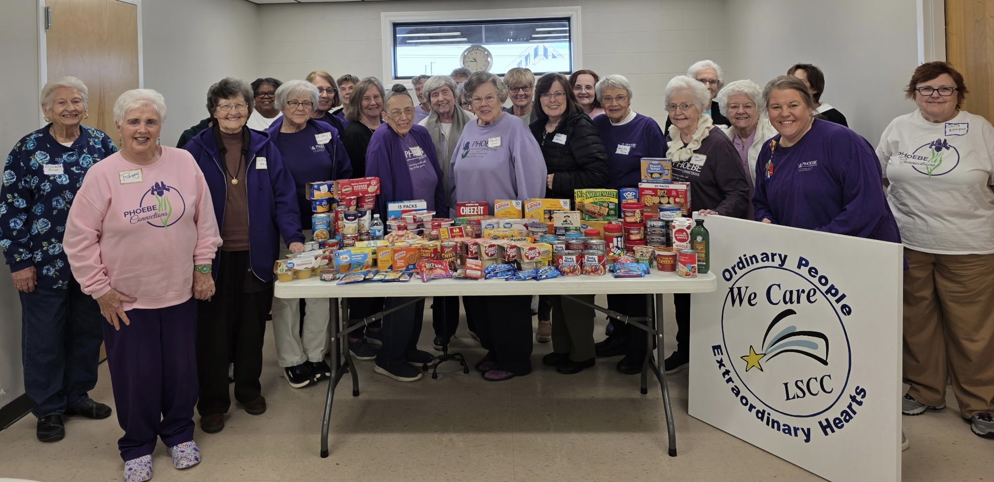 Group of people standing behind a table with food and supplies, in a room with a screen and a banner, smiling.