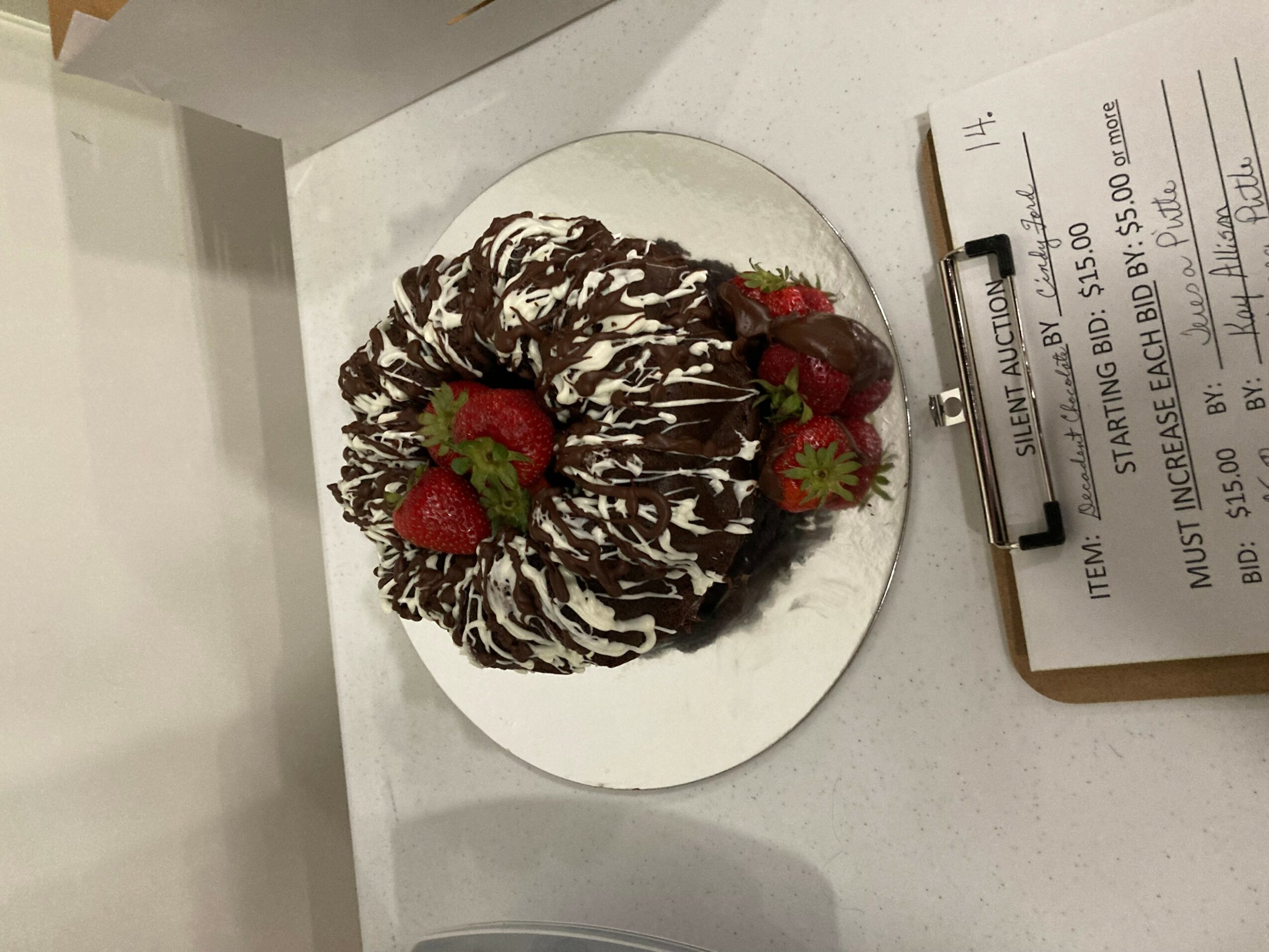 Chocolate-covered dessert with strawberries on a white plate, on a white surface, next to a receipt on a clipboard.