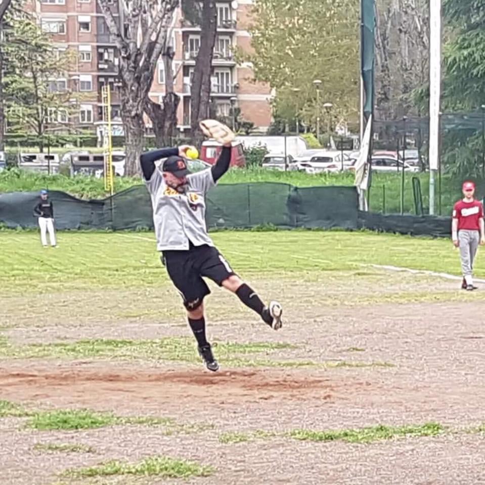 Person jumping on a baseball field with trees and buildings in background, wearing a gray jacket and black pants.