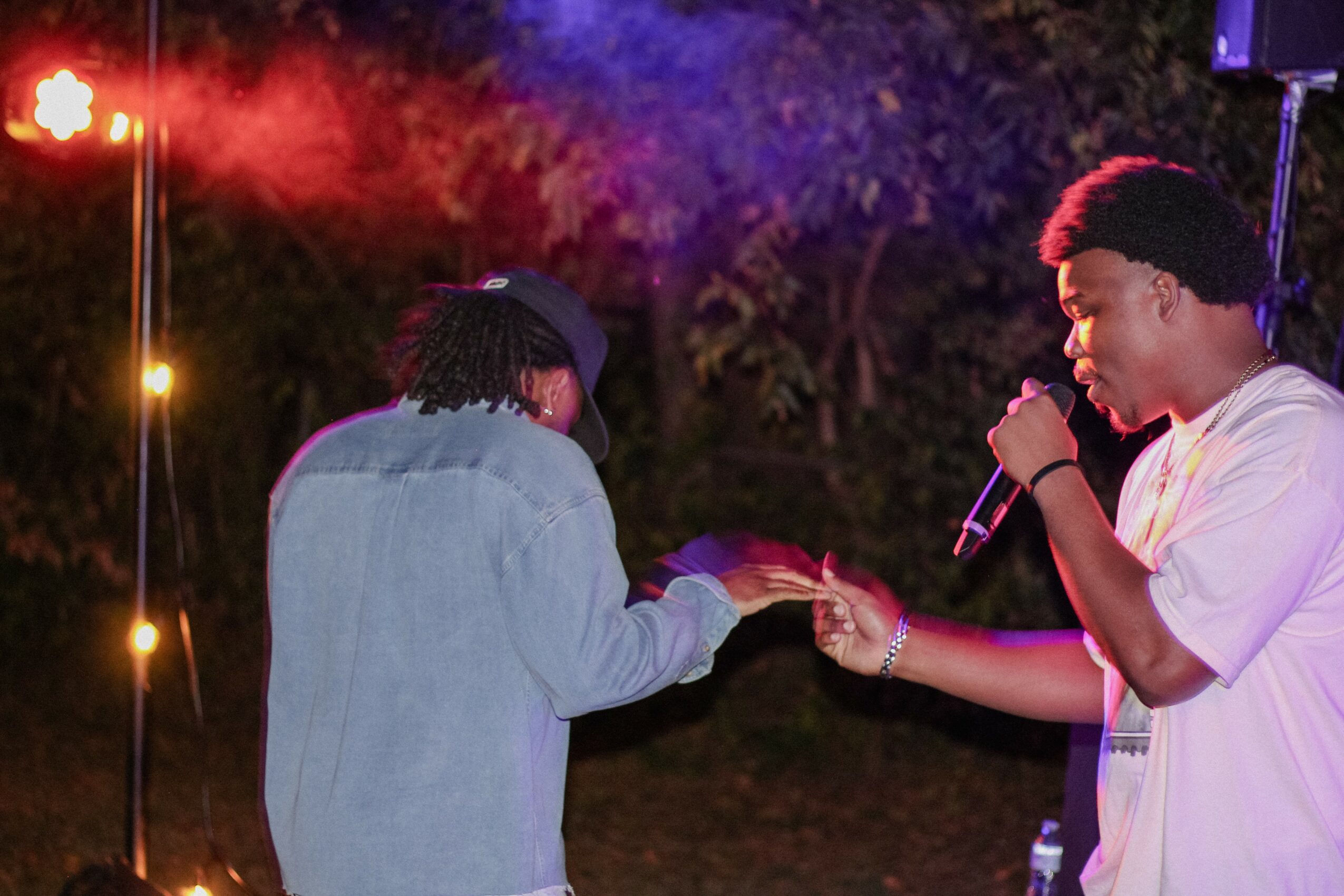 Two people on stage, one holding a microphone, the other reaching out their hand, with colorful lights in the background.