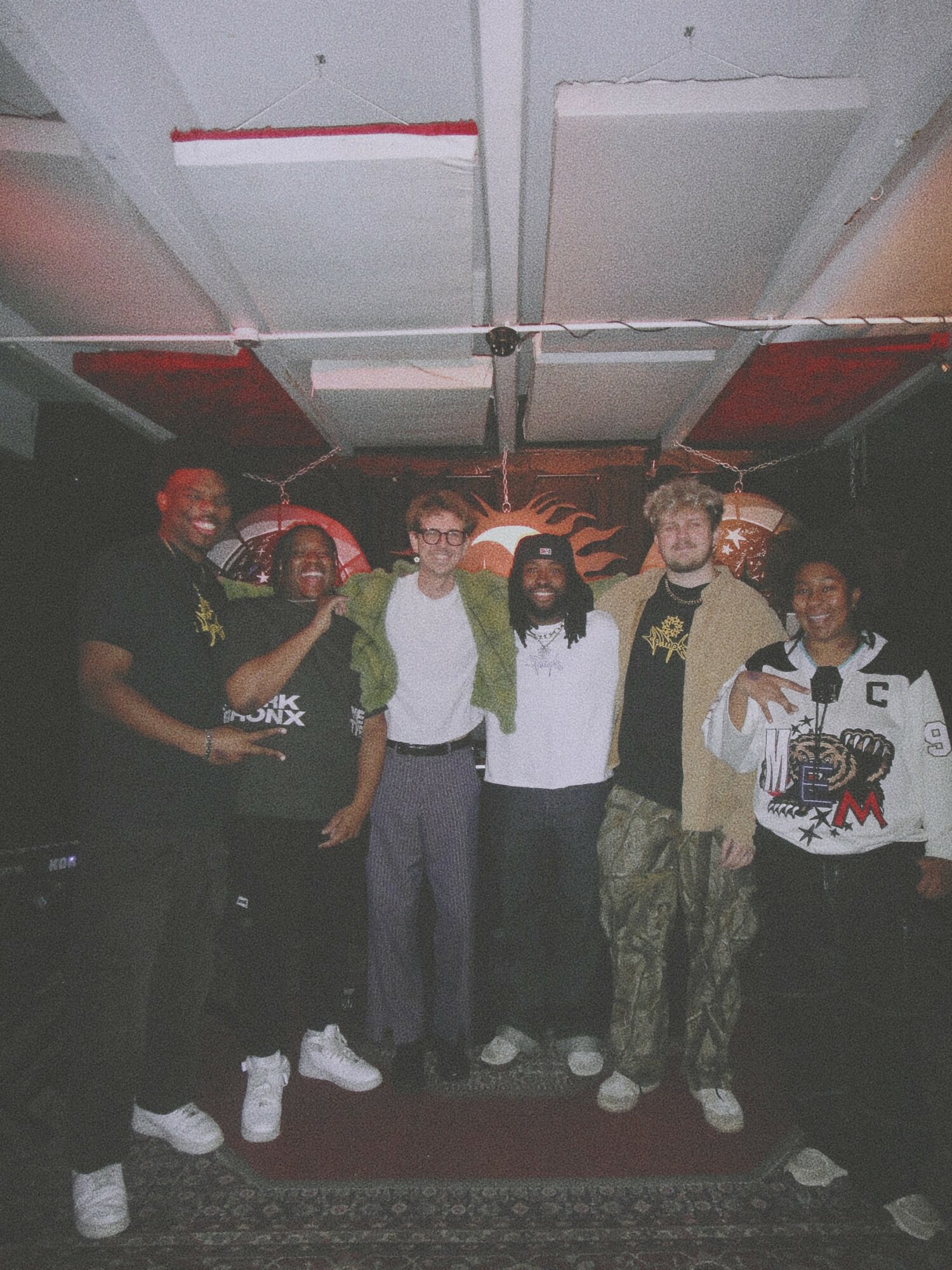 Group of six people standing indoors, some wearing casual clothing and glasses, with a patterned wall behind them.