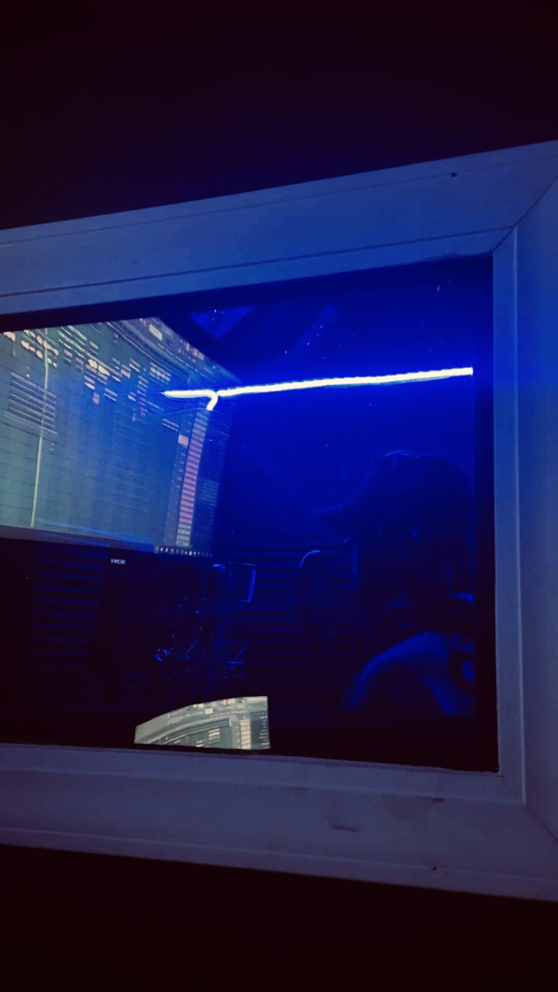 A window with a blue neon light reflecting on the glass, showing a dark room inside with some electronic equipment.