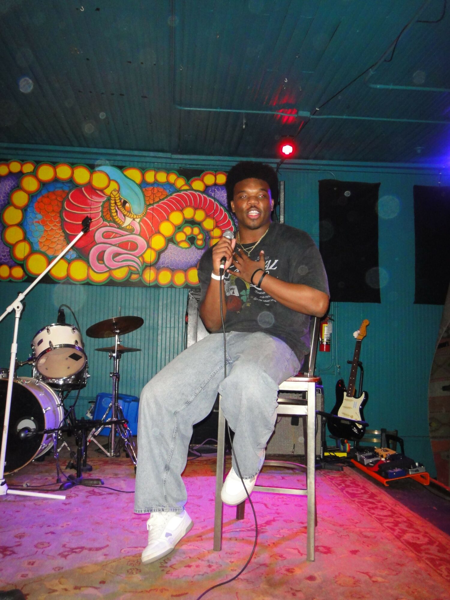 Person singing on stage with musical instruments and colorful backdrop, holding a microphone, wearing casual clothes, in a performance setting.