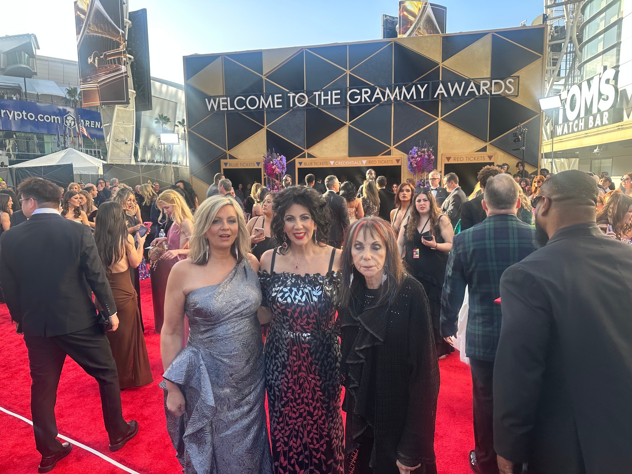 Three women standing on a red carpet at the Grammy Awards, with a large sign and crowd in the background.